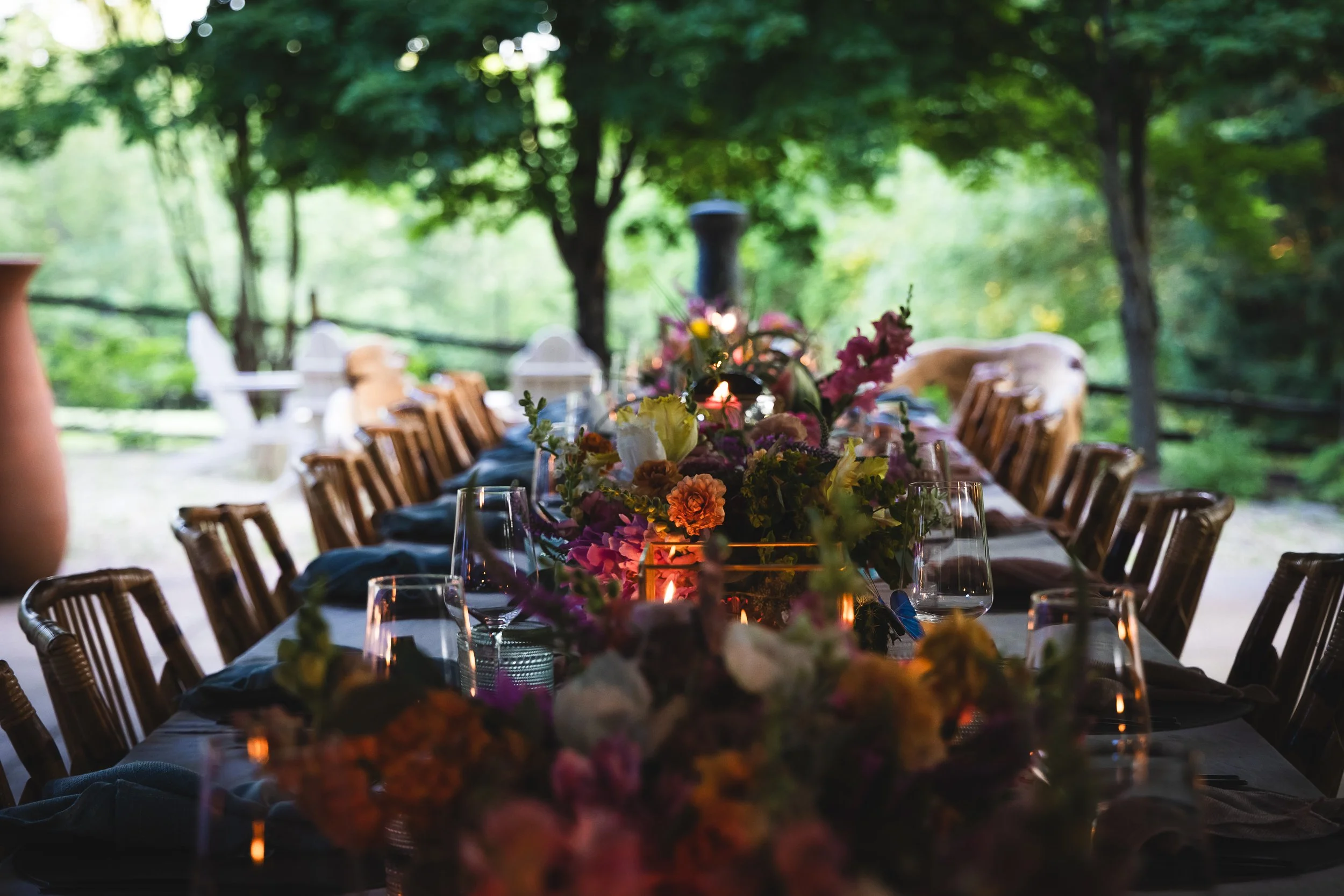 A long outdoor table decorated with colorful flowers and candles, set for a gathering among trees.