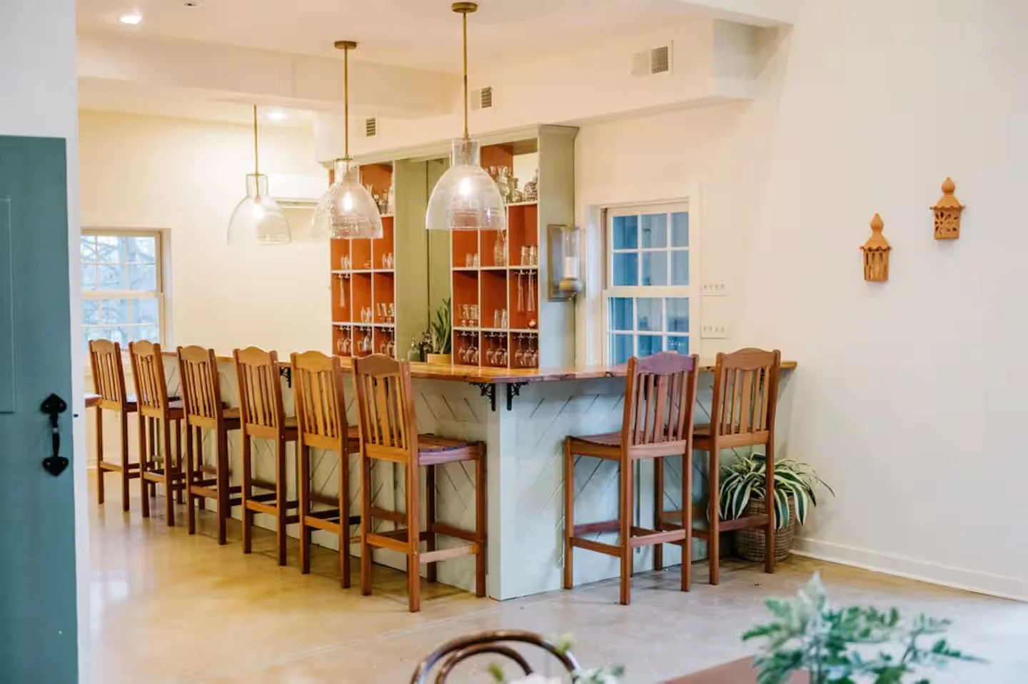 A cozy kitchen with a bar area featuring wooden high chairs, pendant lights, a built-in shelf with glassware, and a small potted plant.