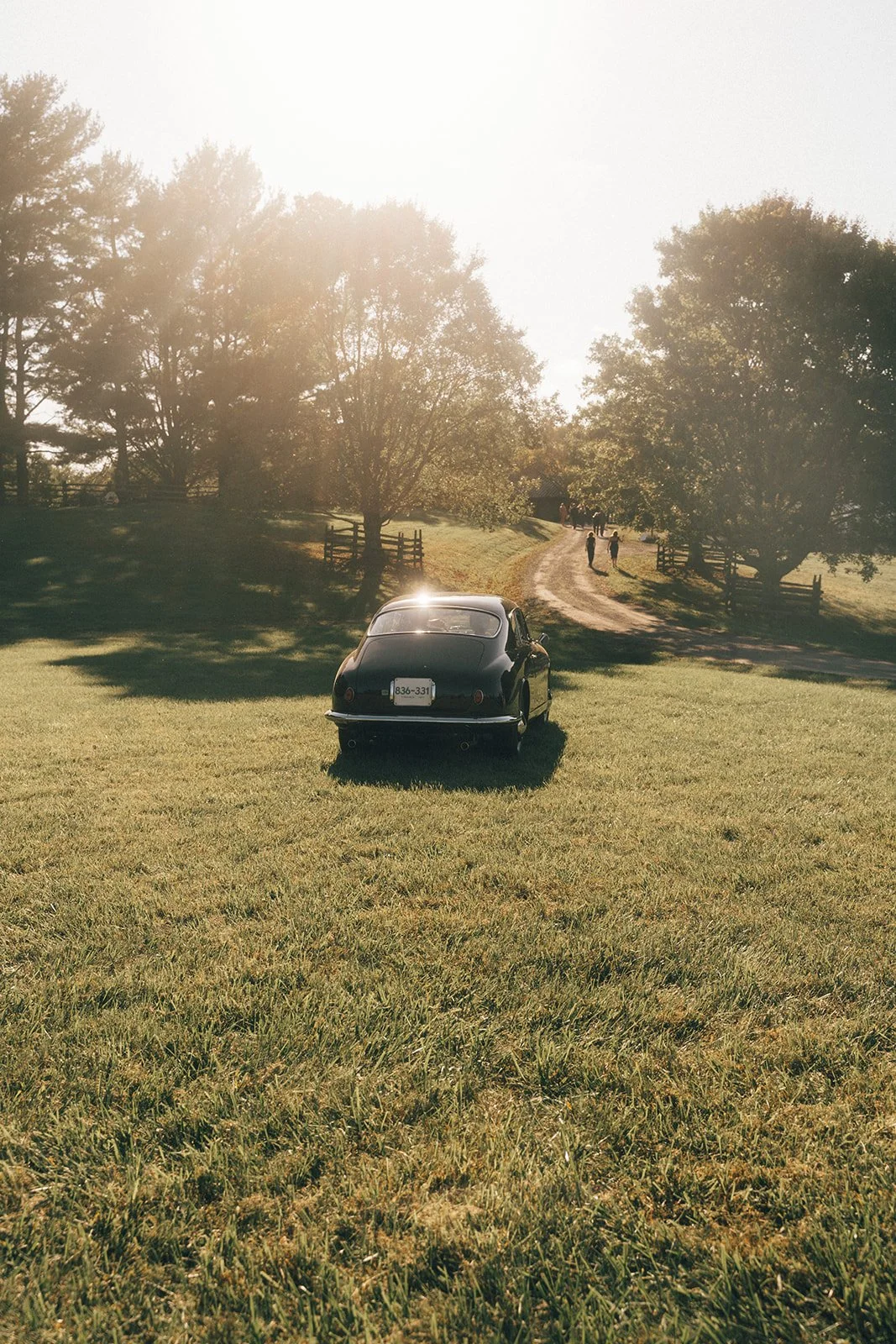 A black vintage car parked on a grassy field with a dirt path and trees in the background on a sunny day.