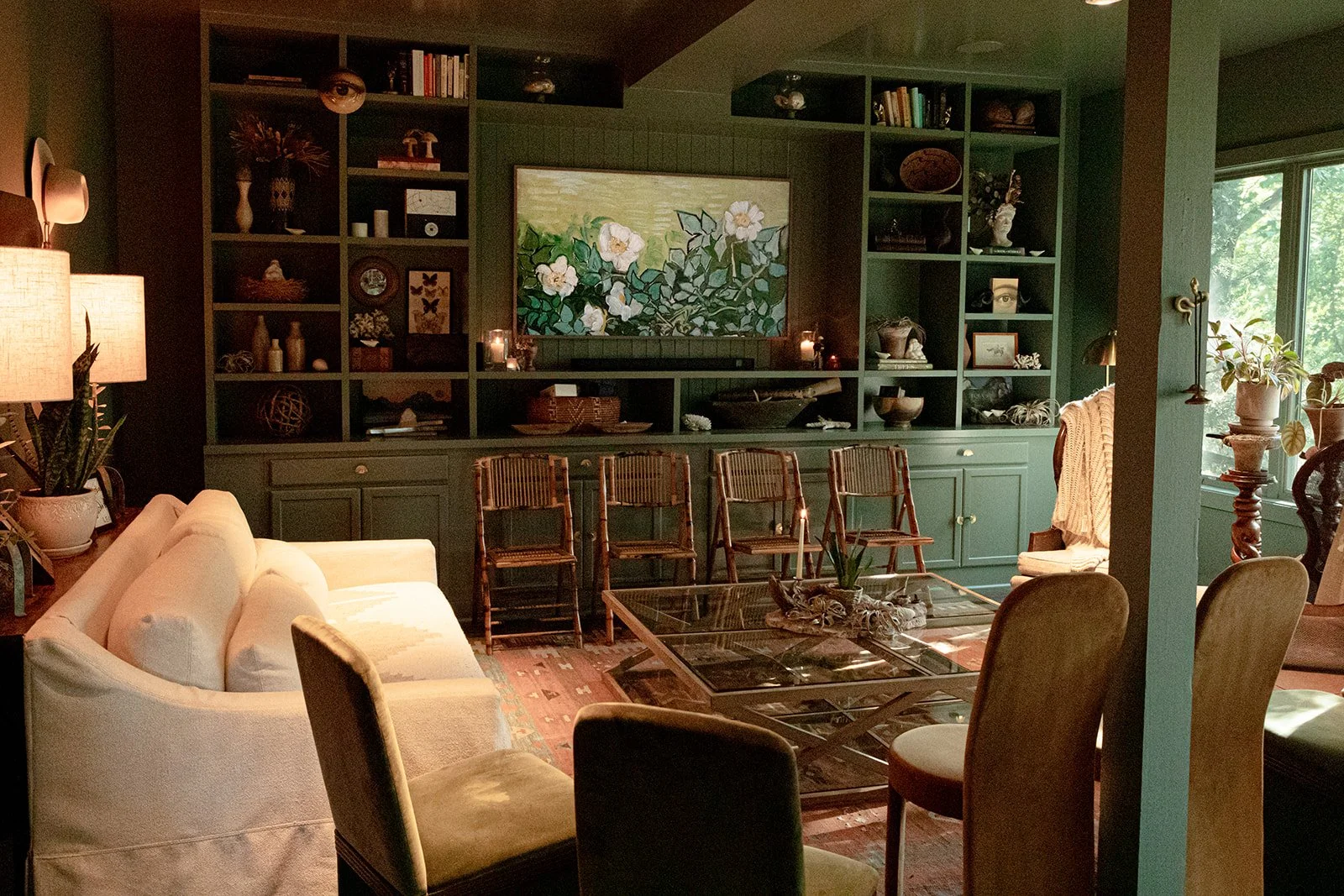 Living room with a green built-in bookshelf filled with decorative items, candles, and books, a wall-mounted painting of white flowers, white sofa, wooden chairs, glass coffee table, and various potted plants.