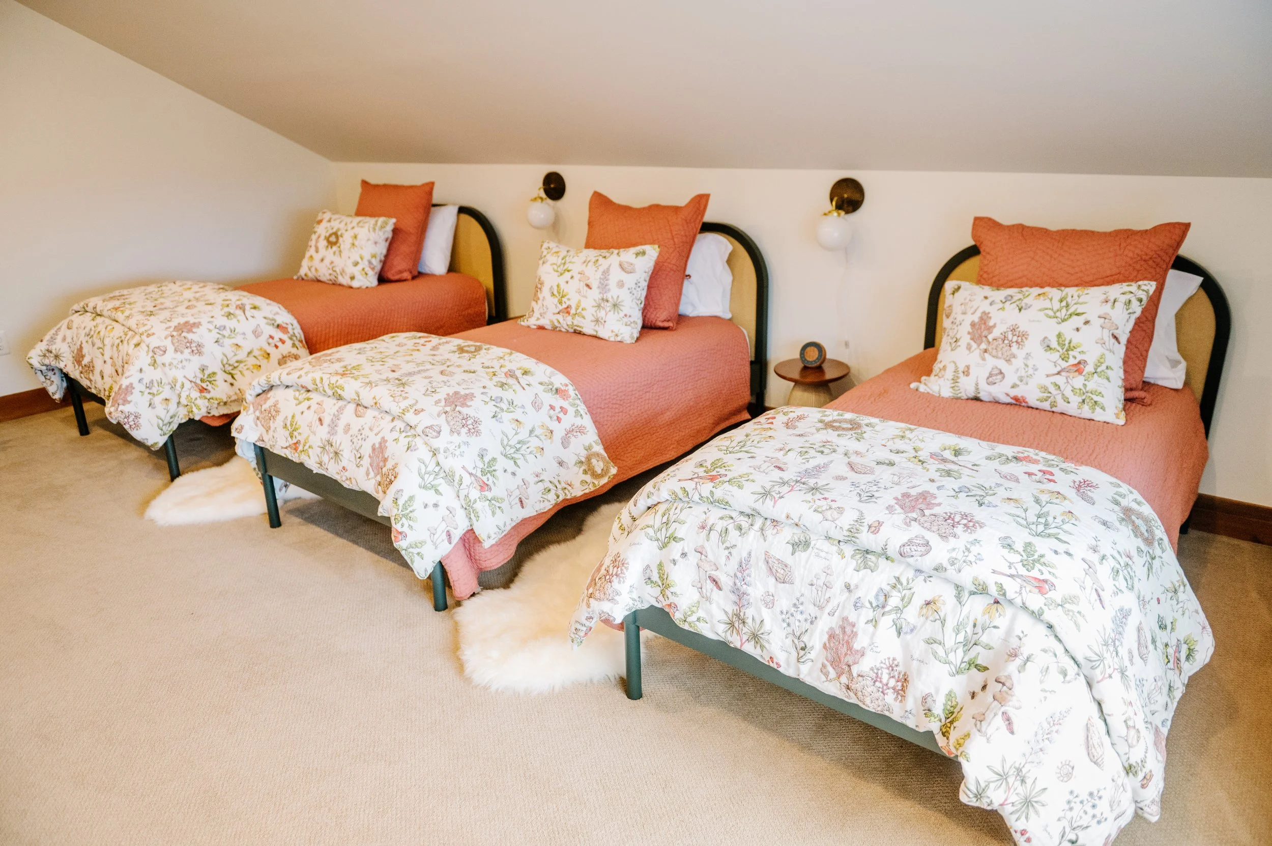 Three beds with matching floral and coral bedding, pillows, and headboards in an attic bedroom. Each bed has a floral pillow, a coral pillow, and is flanked by wall-mounted lamps and a small wooden nightstand.