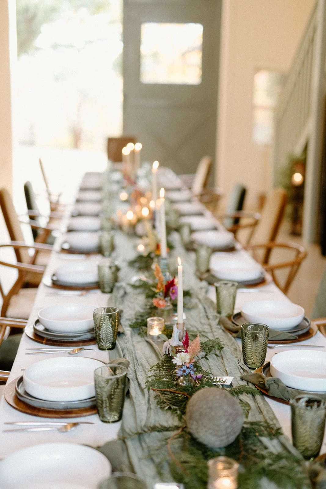 A long dining table set for a special occasion with white plates, dark green glasses, silverware, lit candles, and floral centerpieces with greenery and colorful flowers, in a bright room with natural light.