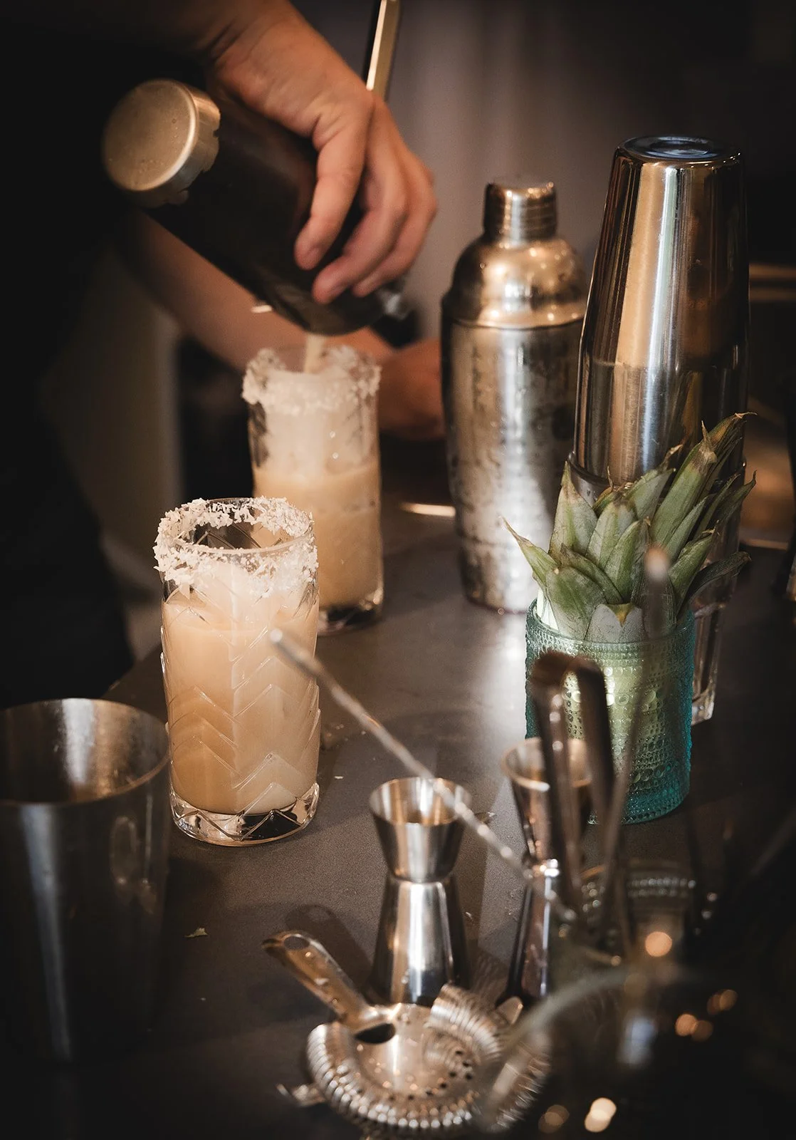 A bartender preparing cocktails with a shaker, two drinks with salted rims, a bar spoon, a jigger, a cocktail strainer, pineapple leaves, and bar tools on the counter.