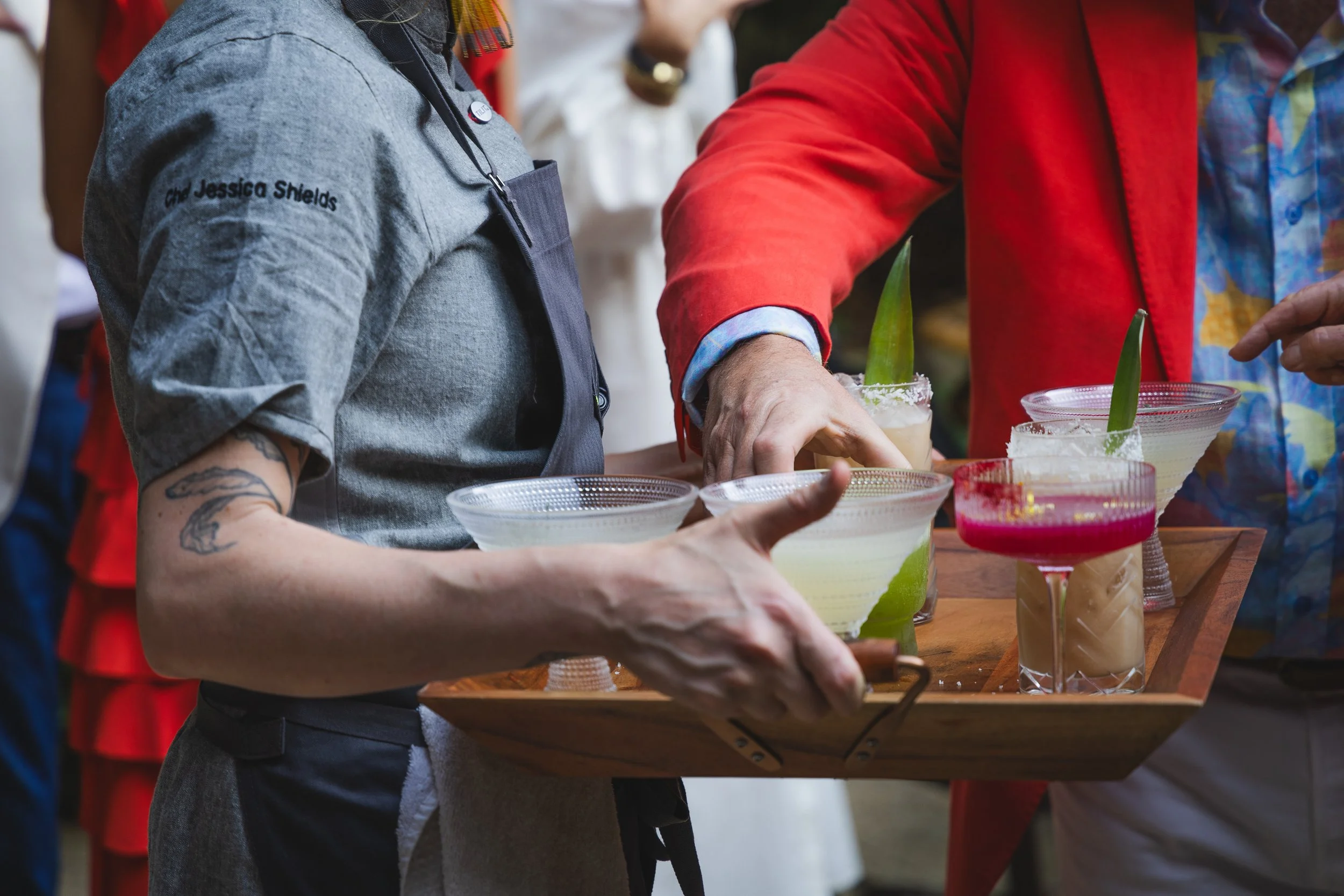People serving and holding various colorful cocktails on a wooden tray during a social gathering or event.