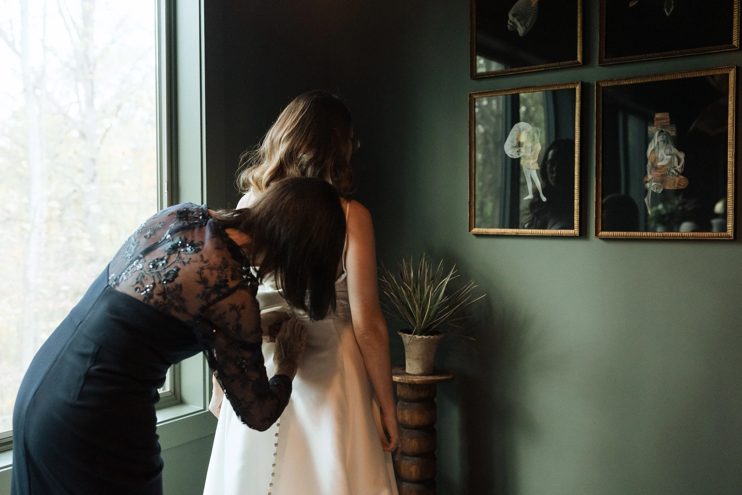 A woman helping another woman prepare for an event, standing near a window with a green wall and framed artwork on the wall.