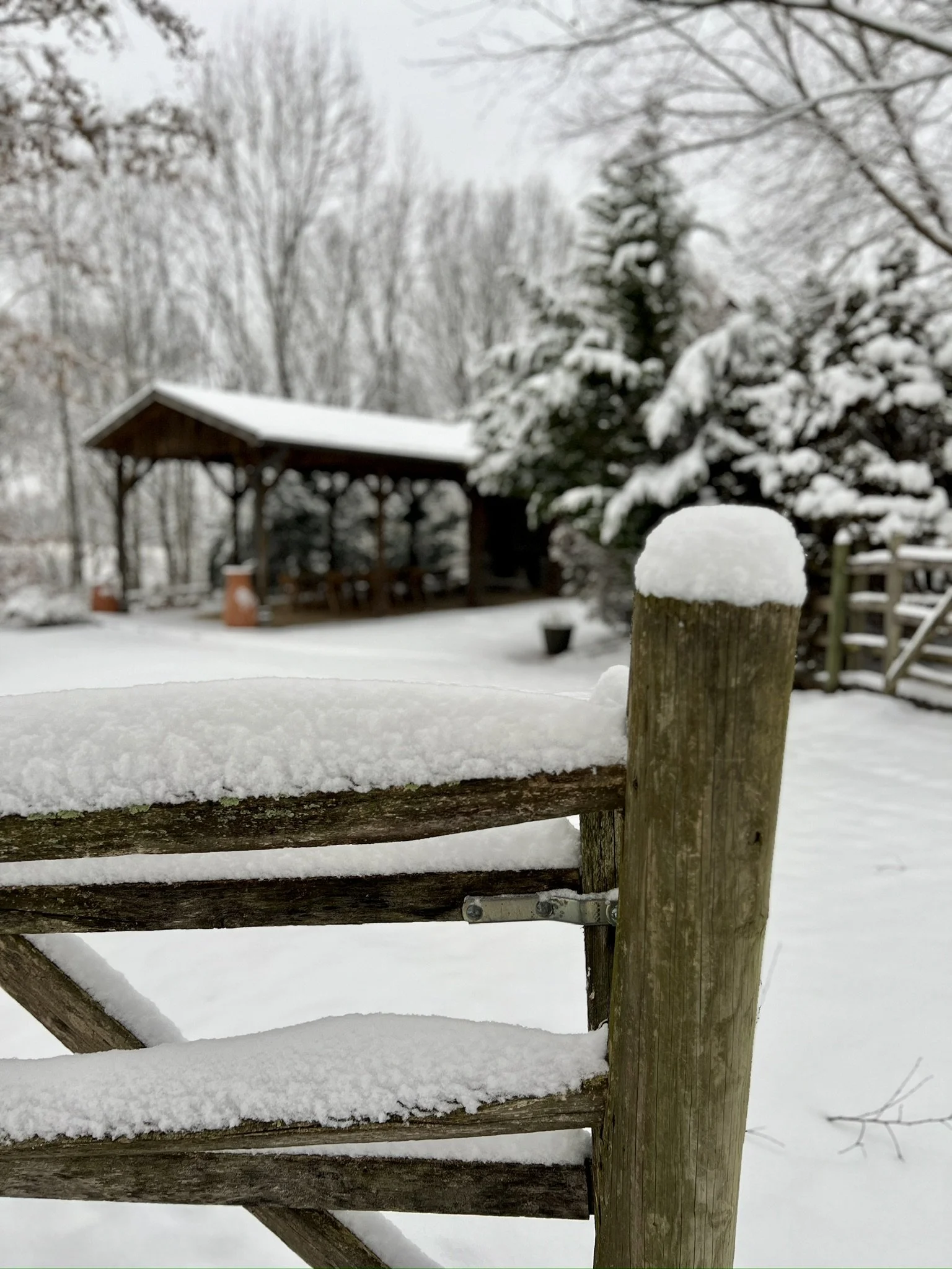 Snow-covered wooden fence in a snowy outdoor setting with a pavilion and trees in the background.