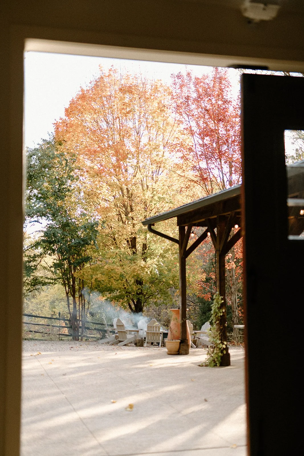 View of a backyard patio with fall-colored trees, outdoor seating, and a faint plume of smoke.