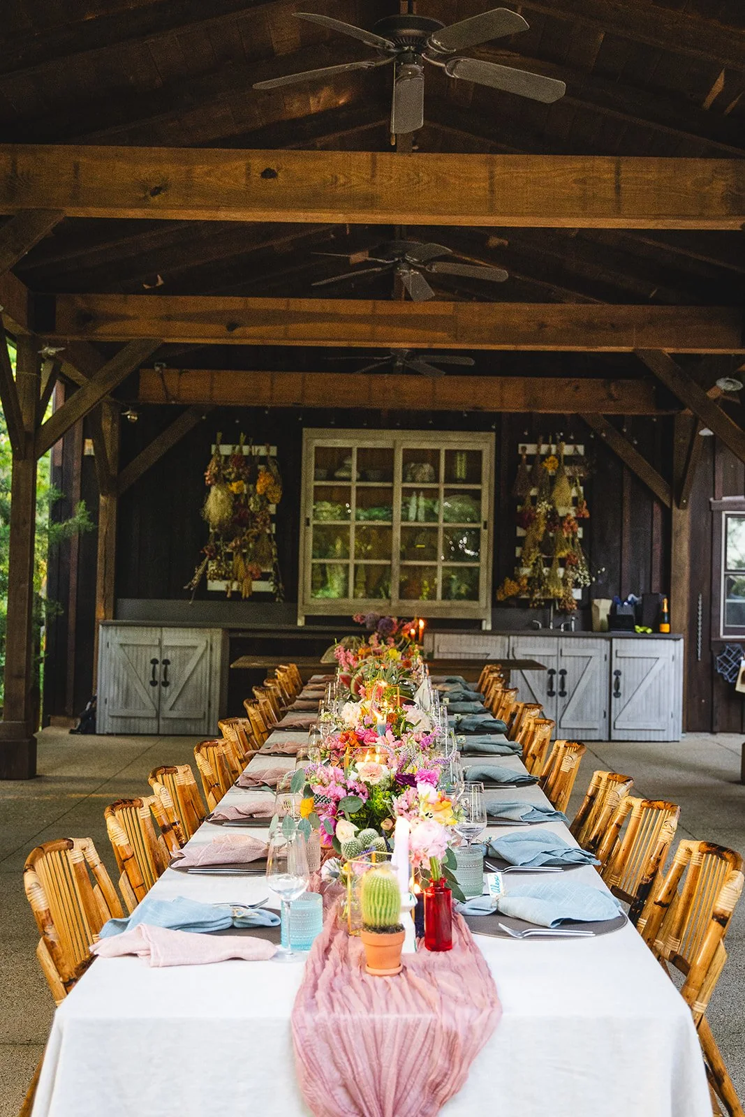 A long rectangular dining table decorated with pink, purple, and yellow flowers, greenery, candles, and place settings, set for a celebration inside a rustic open-air wooden pavilion with ceiling fans.