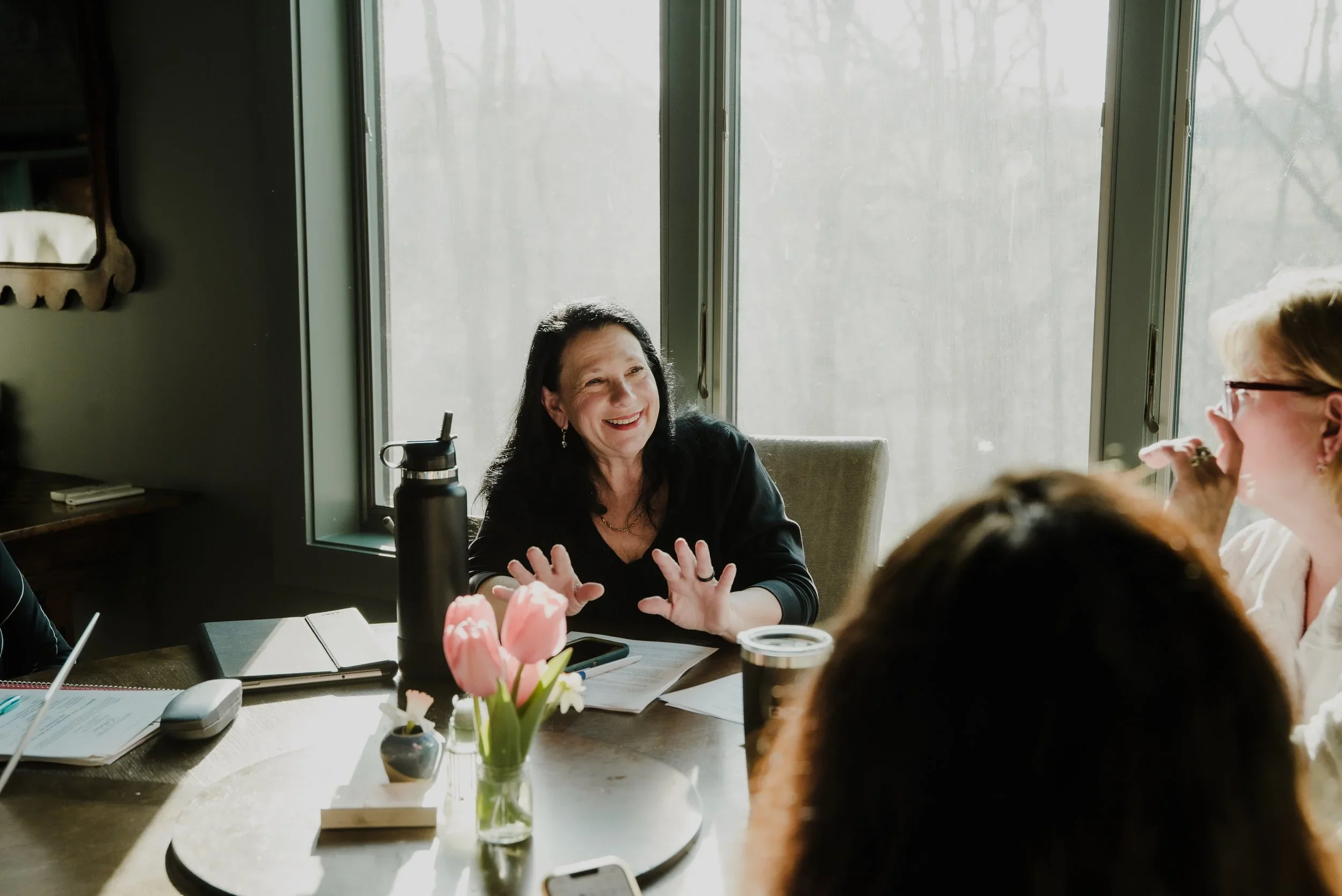 Women sitting at a table having a conversation, with one woman smiling and gesturing, flowers and notebooks on the table.