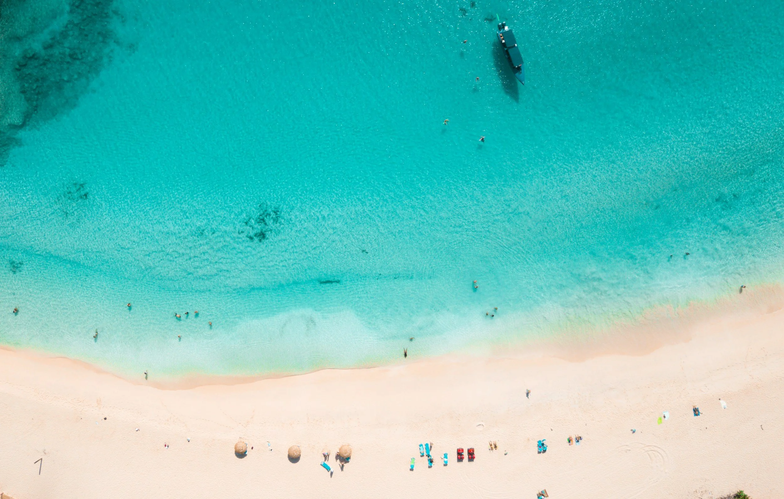 Aerial view of a beach with turquoise water, white sand, and people swimming, sunbathing, and walking along the shore.