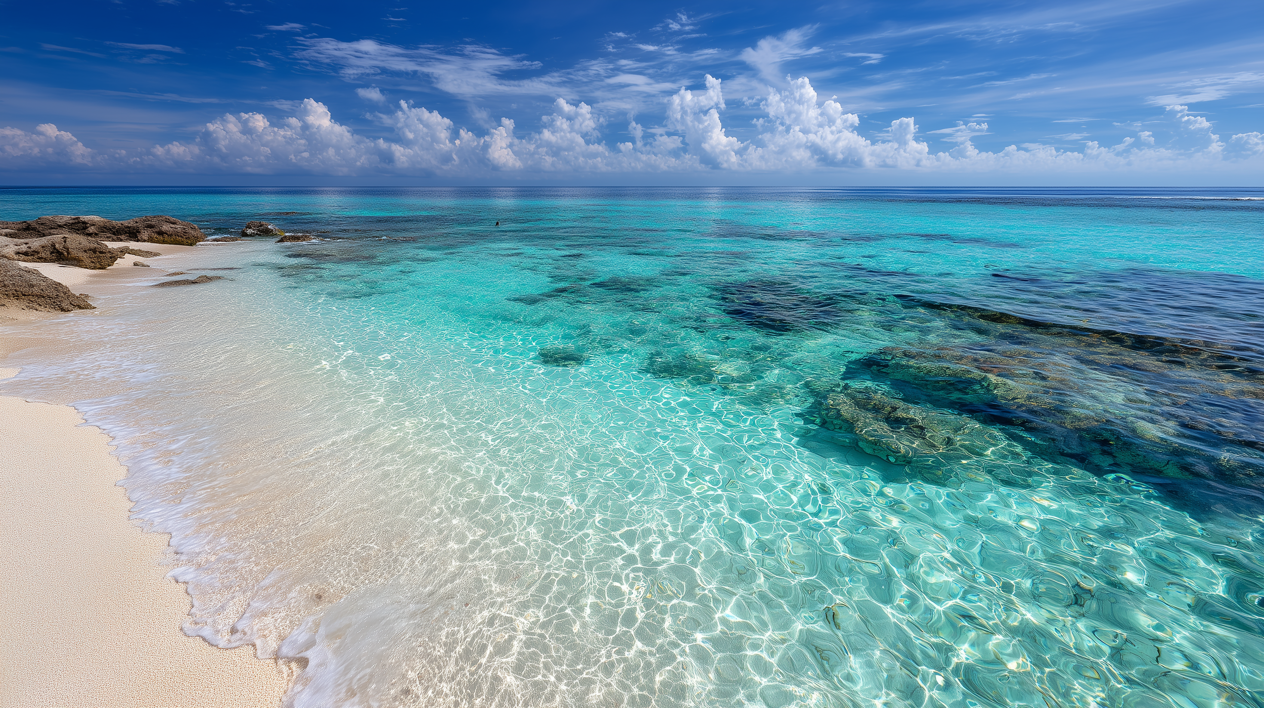 Clear turquoise water lapping onto a sandy beach with rocks, under a blue sky with fluffy clouds