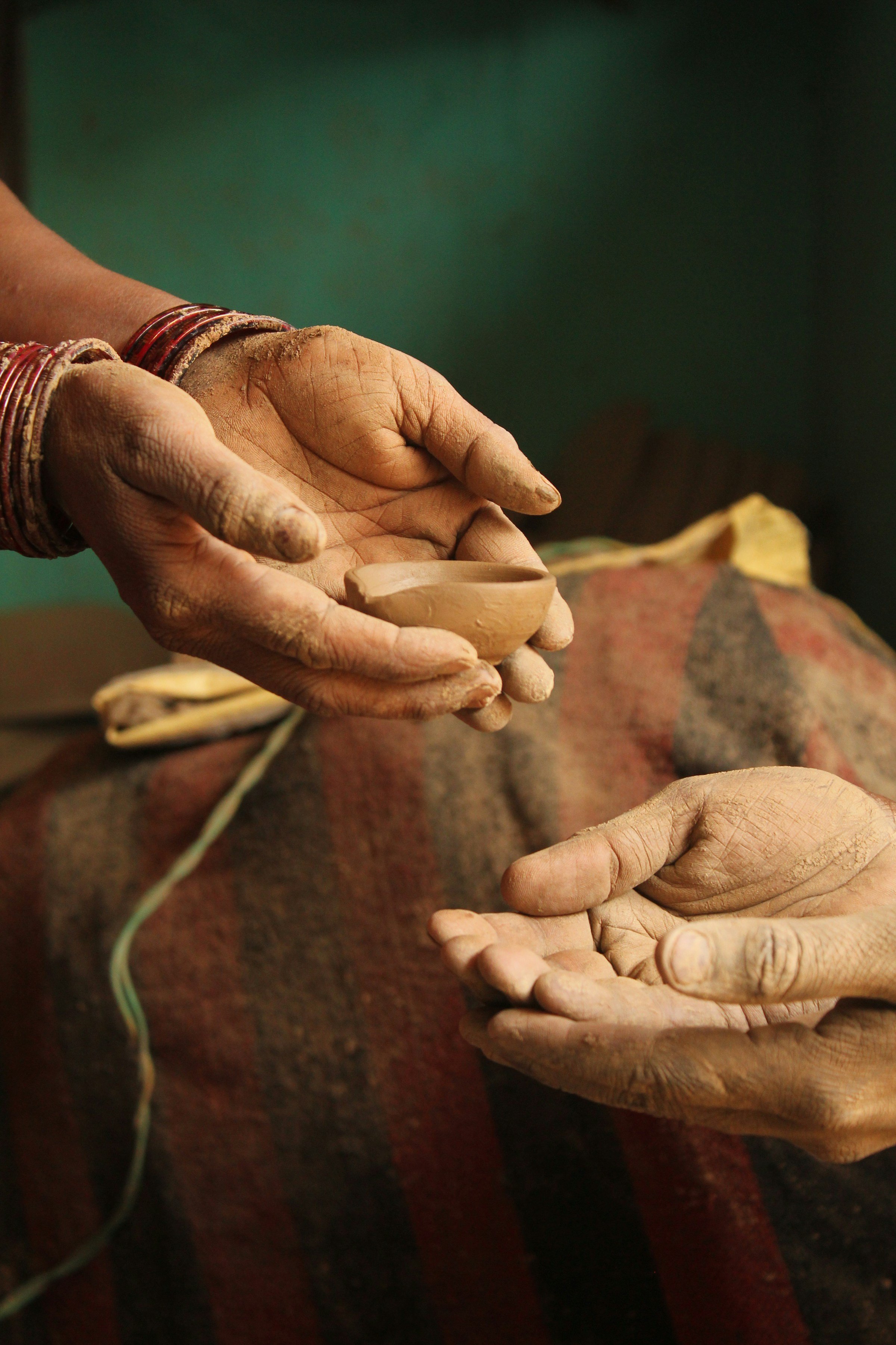 A person with weathered hands and multiple red bangles on their wrist is working with clay, shaping a small bowl in their hands. The background is blurred with warm earthy tones.