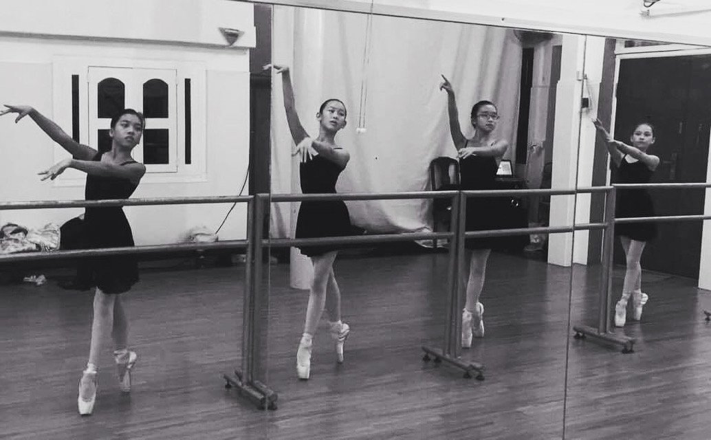 Four ballet dancers in black leotards and tutus practicing at a dance studio, standing at the barre with arms raised.