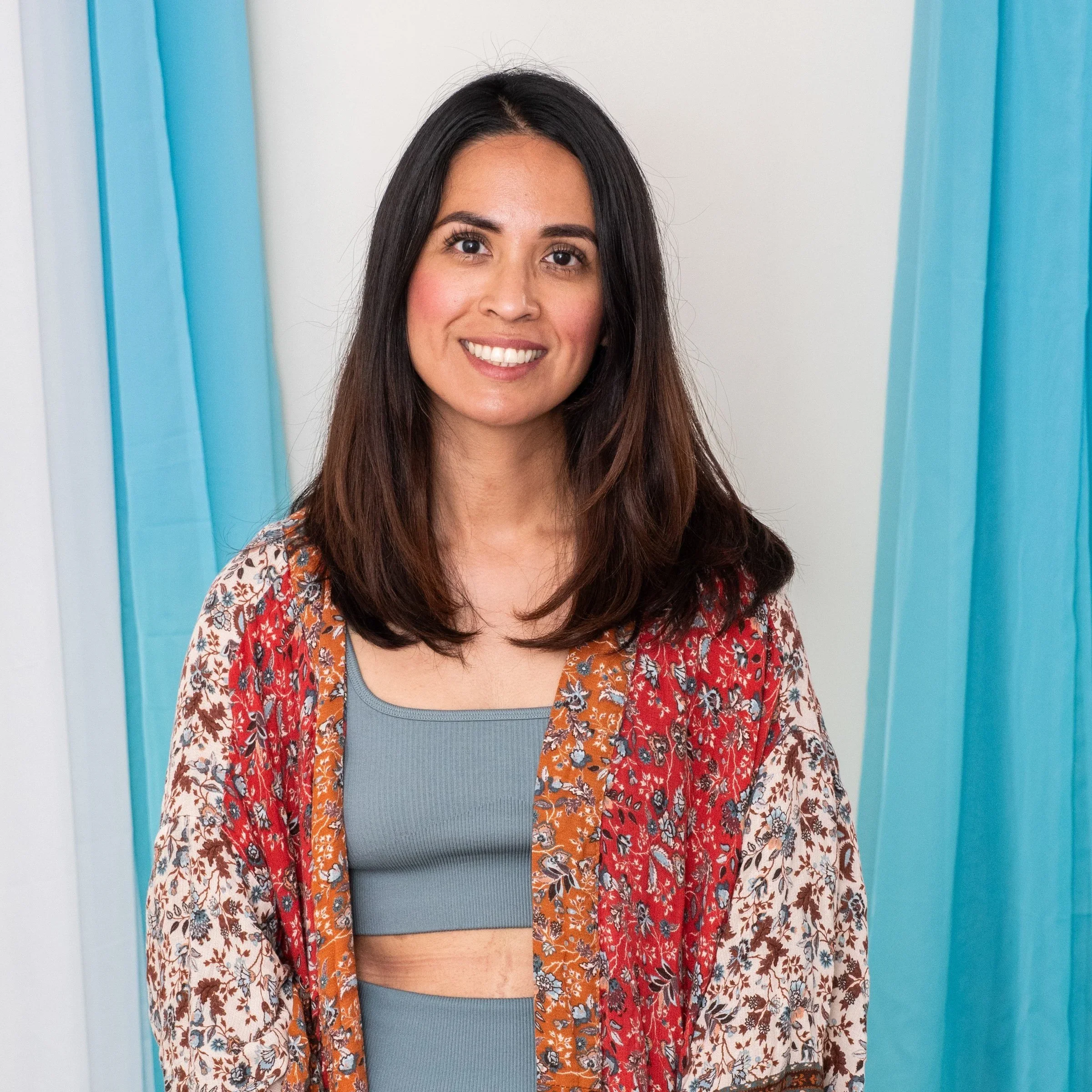 Smiling woman with long dark hair wearing a blue top and a colorful, patterned kimono-style cover-up, standing in front of a white and blue background.