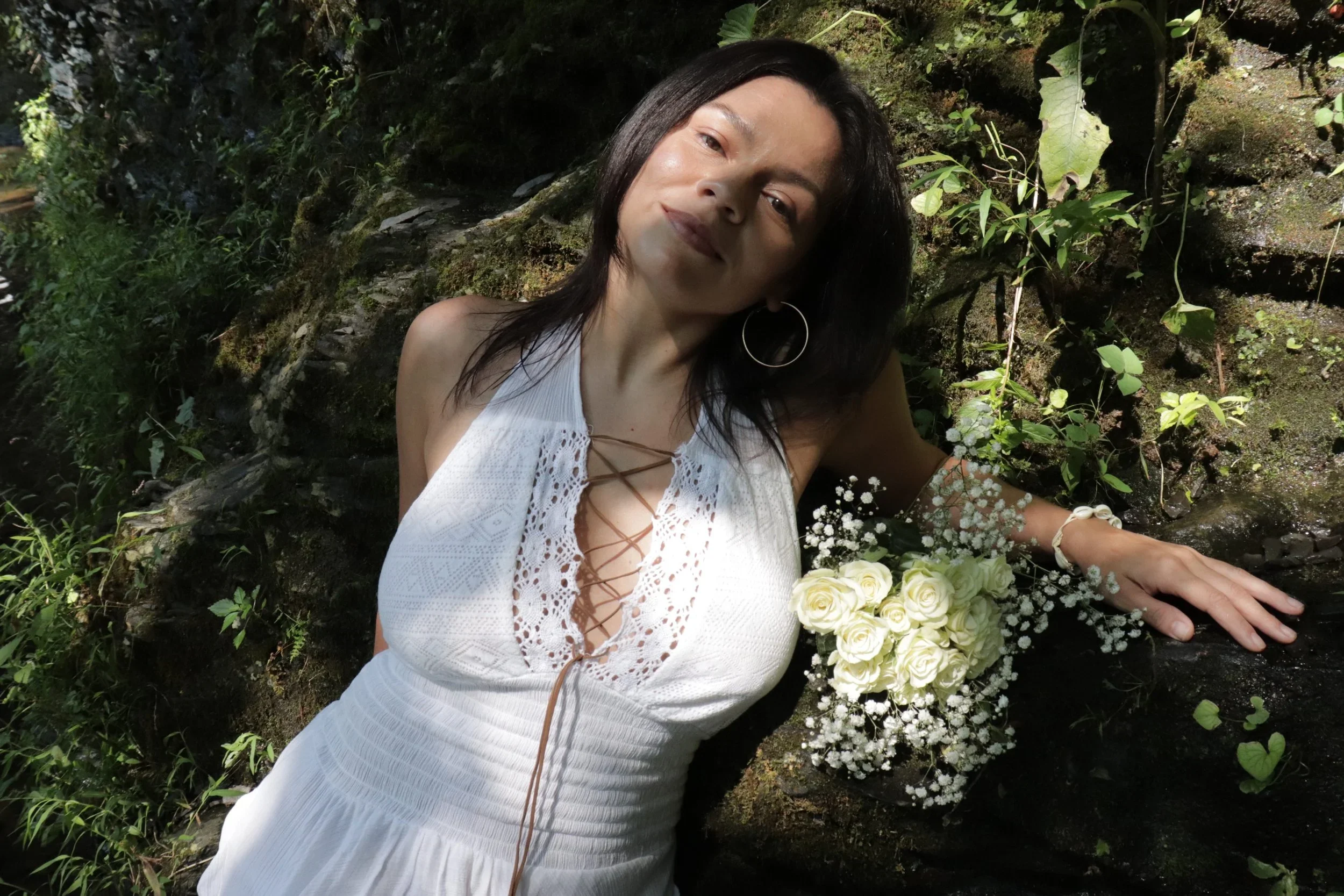 A woman in a white dress with lace details and a plunging neckline, wearing hoop earrings, lying on a mossy rock surrounded by greenery, with a bouquet of white roses and baby's breath flowers resting on the rock next to her.
