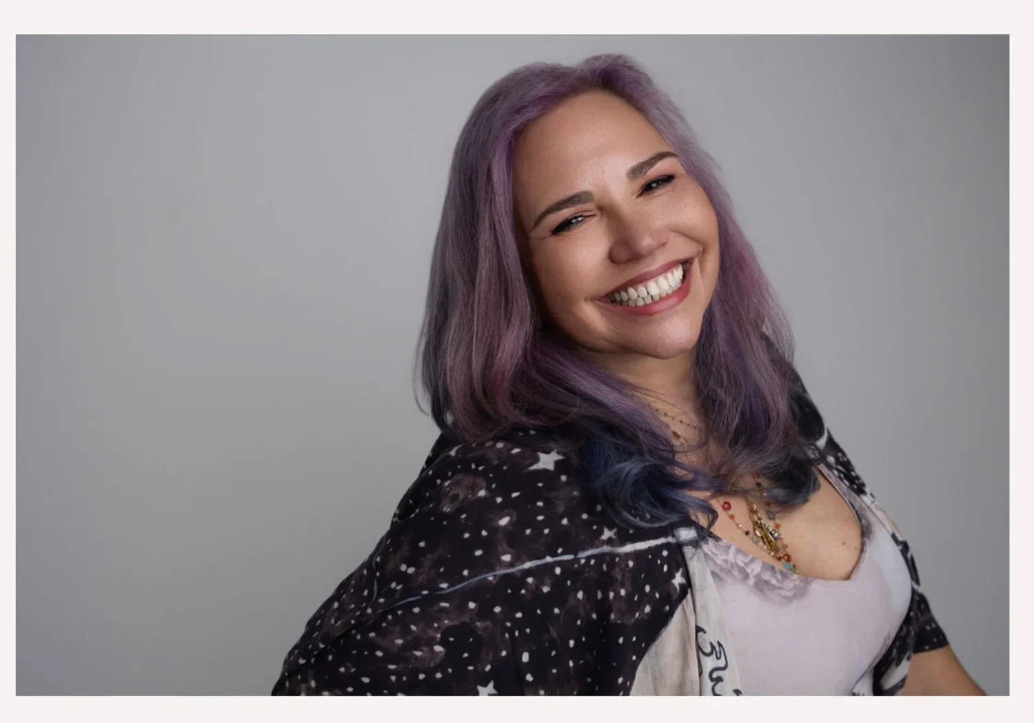 Smiling woman with purple and blue hair, wearing a black patterned jacket and layered necklaces, against a plain gray background.