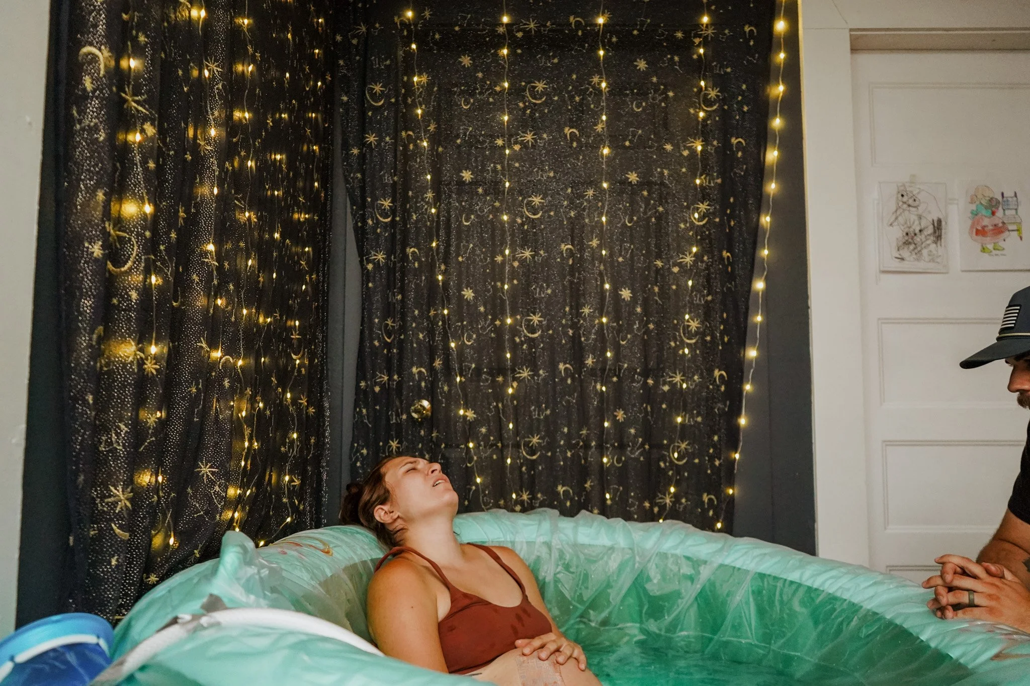 A woman in a brown tank top in a birth pool, smiling with her head tilted back, while someone stands nearby. The background features black curtains decorated with gold moon and star patterns, with string lights hanging down.