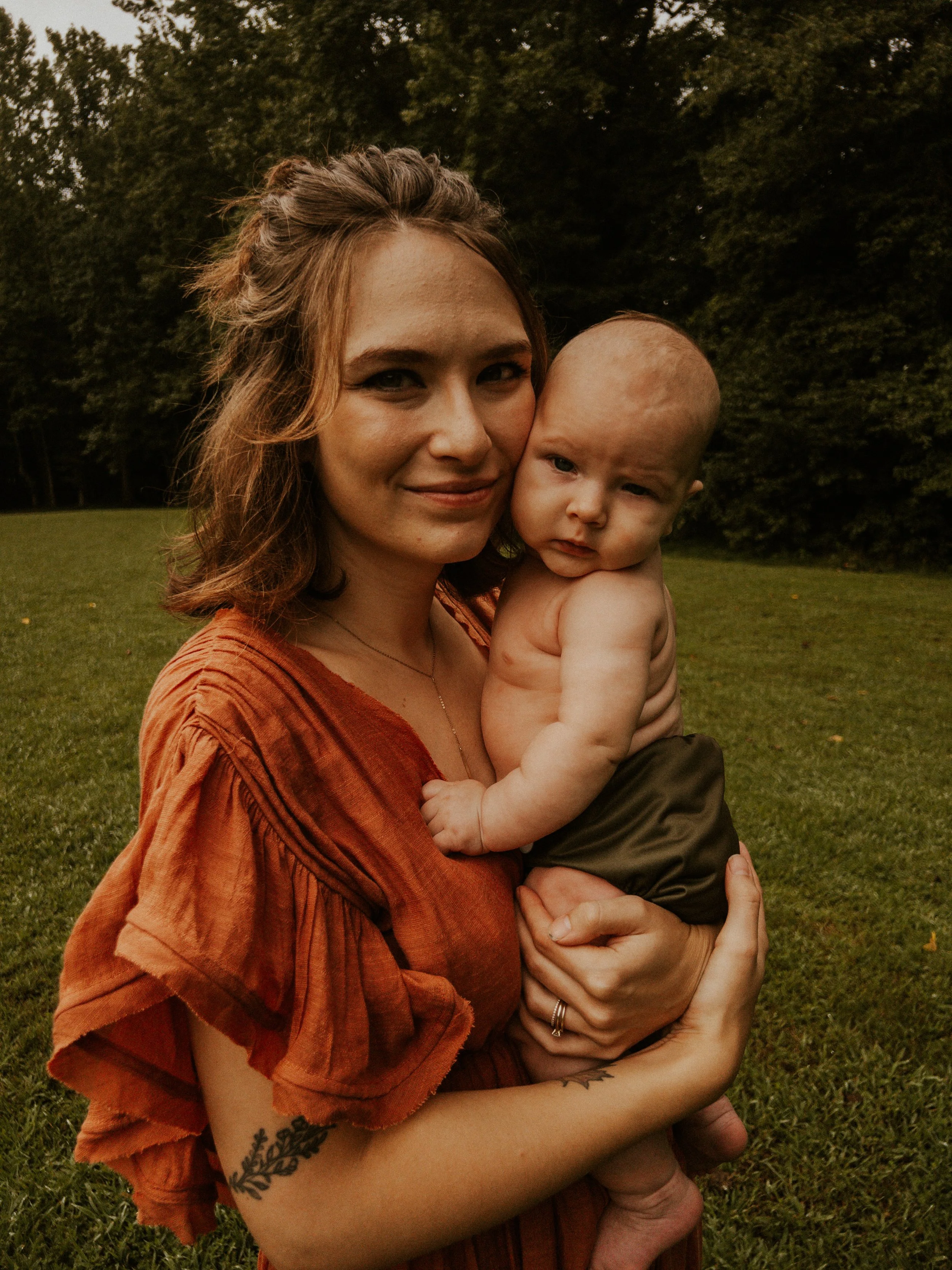 A woman holding a baby outdoors on grass, with trees in the background. The woman has light skin, long wavy hair, and is wearing an orange shirt. The baby is shirtless, with light skin, a bald head, and is making a slightly confused expression.