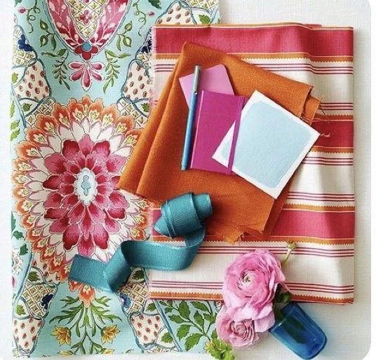 Flat lay of fabric, notebooks, a pen, a blue ribbon, pink flowers, and a small blue vase.