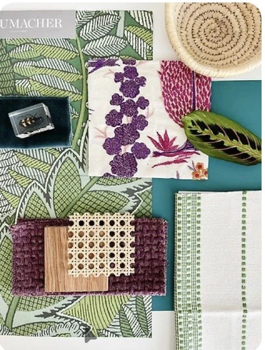 A flat lay of various textiles and decorative items including patterned fabric, a woven basket, a small wooden and lace square, a green and black leaf, a jewelry box with rings, and a book titled 'UMACHER', arranged on a colorful Botanical print clot