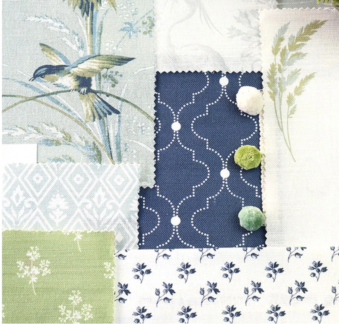 A flat lay of various fabric swatches with floral and bird patterns in green, white, navy blue, and beige, with three pom-poms in white, green, and light green placed on a navy blue swatch.