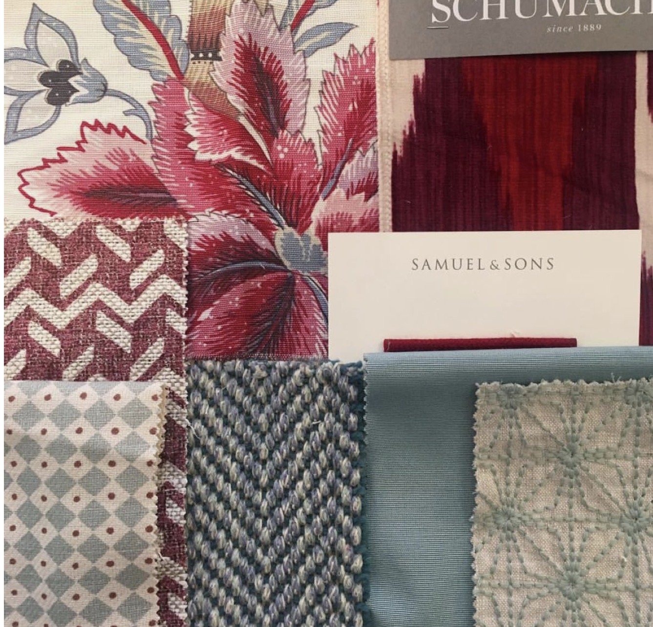 A collection of fabric swatches featuring various patterns and textures, including floral, geometric, and textured designs, along with color samples in shades of red, blue, beige, and cream, arranged on a surface with a sample card from Samuel & Sons