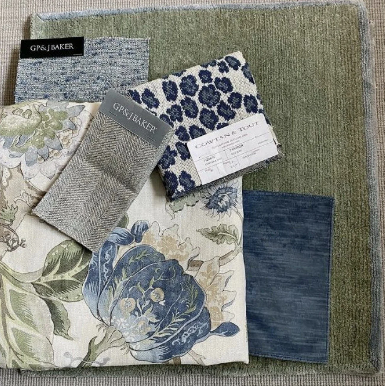 Several fabric swatches with various patterns and textures laid out on a green textured surface. Some swatches have labels, including one from GP&J Baker and another from Coutan & Tout, featuring floral and geometric designs in shades of blue, green,
