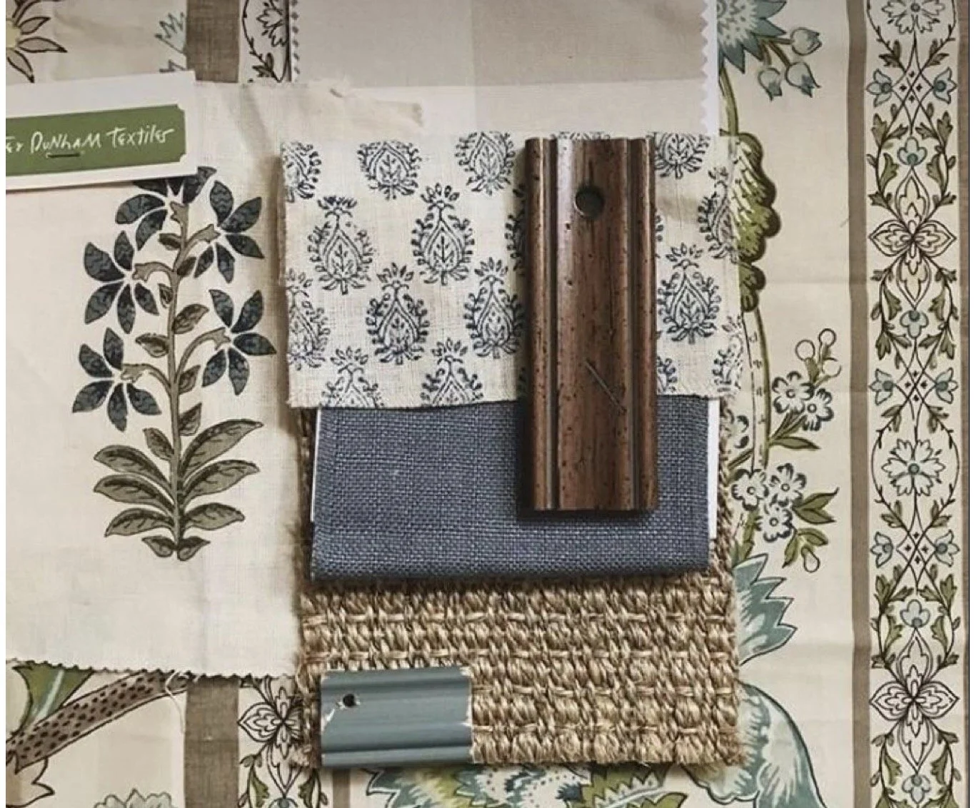 A collection of fabric and material samples arranged on a patterned floral tablecloth, including a piece of black and white printed fabric, a raffia square, a black mesh fabric, a piece of blue material, a metal picture rail hook, and a wooden dowel.