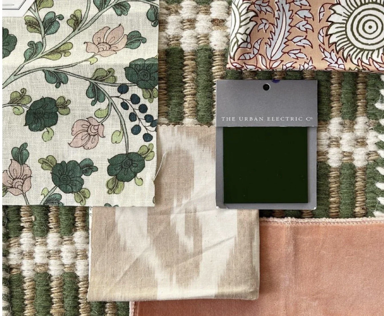 Collection of fabric swatches with floral, patterned, and solid designs, arranged on a textured woven surface, with a small blackboard from The Urban Electric Co. placed among them.