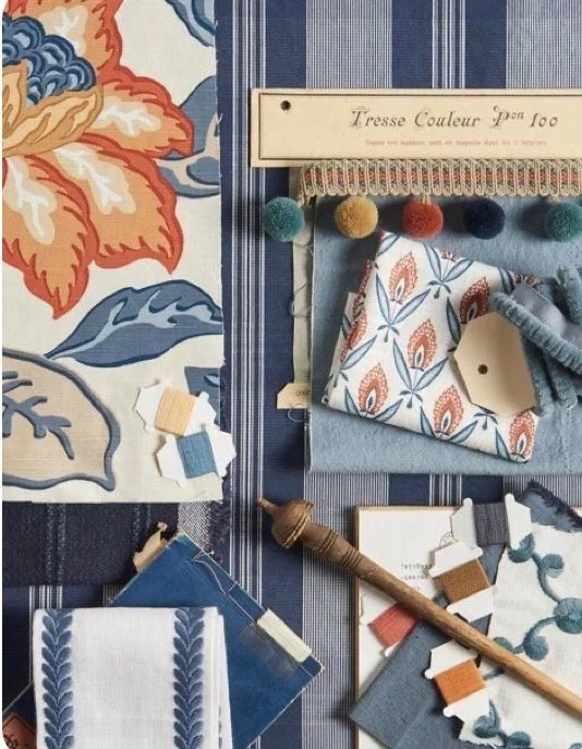 A fabric swatch display with blue, orange, and white patterns, wool yarns in various colors, a fabric comb, a wooden spinning wheel, and sample cards on a striped and floral fabric background, used for textile and sewing projects.