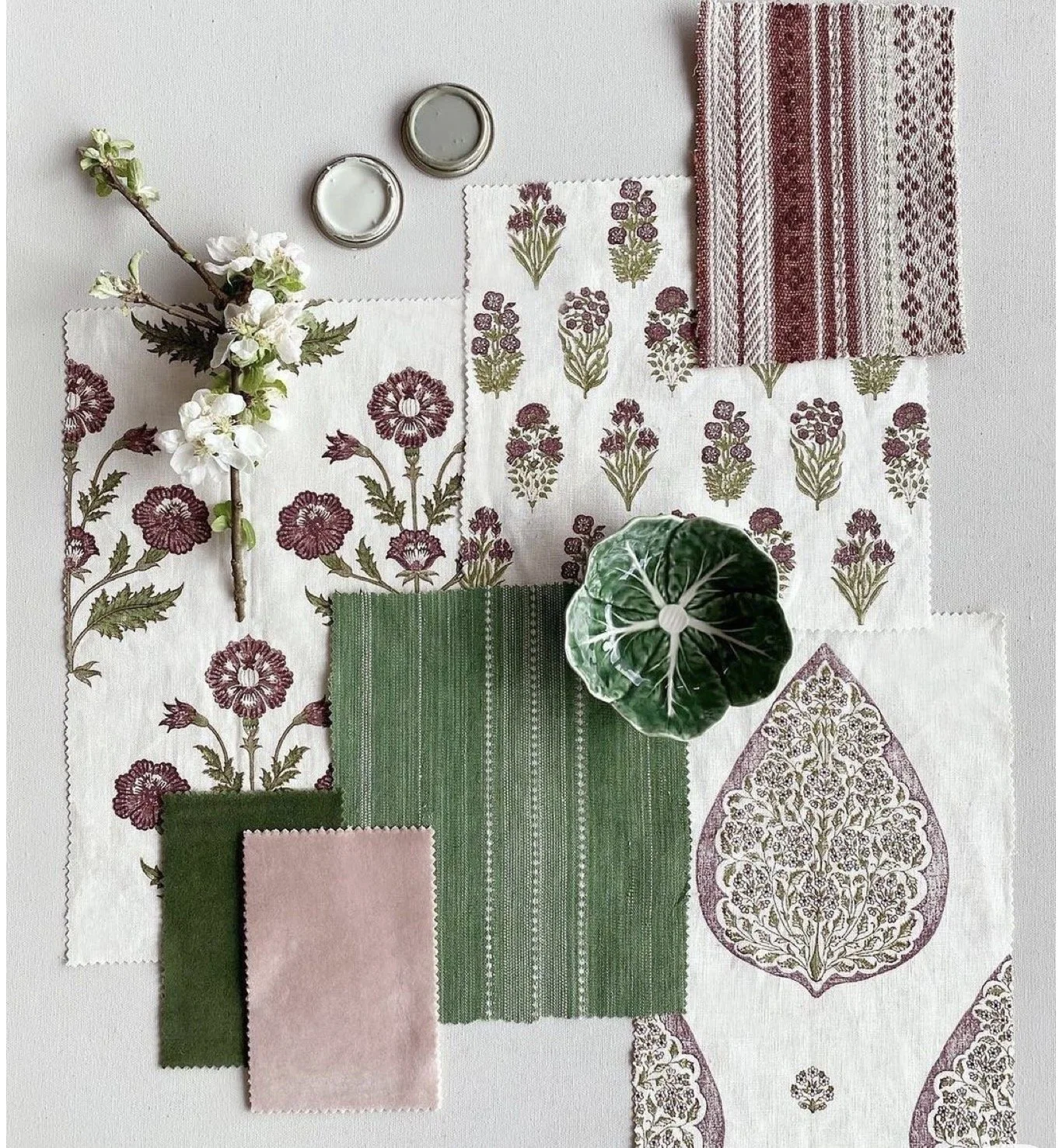 A collection of fabric swatches with floral and geometric patterns, a small branch with white flowers, a small green plant, and two small metal containers on a white surface.