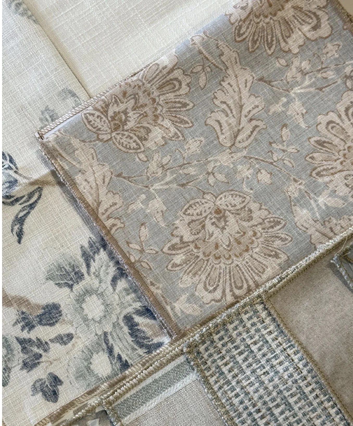 Close-up of folded patterned fabric, highlighting intricate floral and leaf designs in beige, brown, and light blue tones, with a textured border.