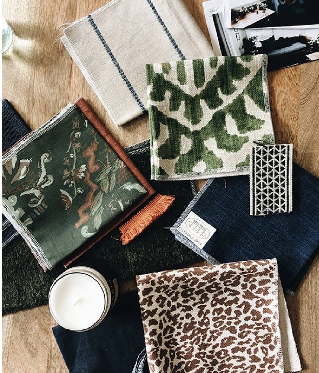 A collection of assorted fabric swatches with different patterns and textures, a lit white candle, and some magazines, all arranged on a wooden surface.