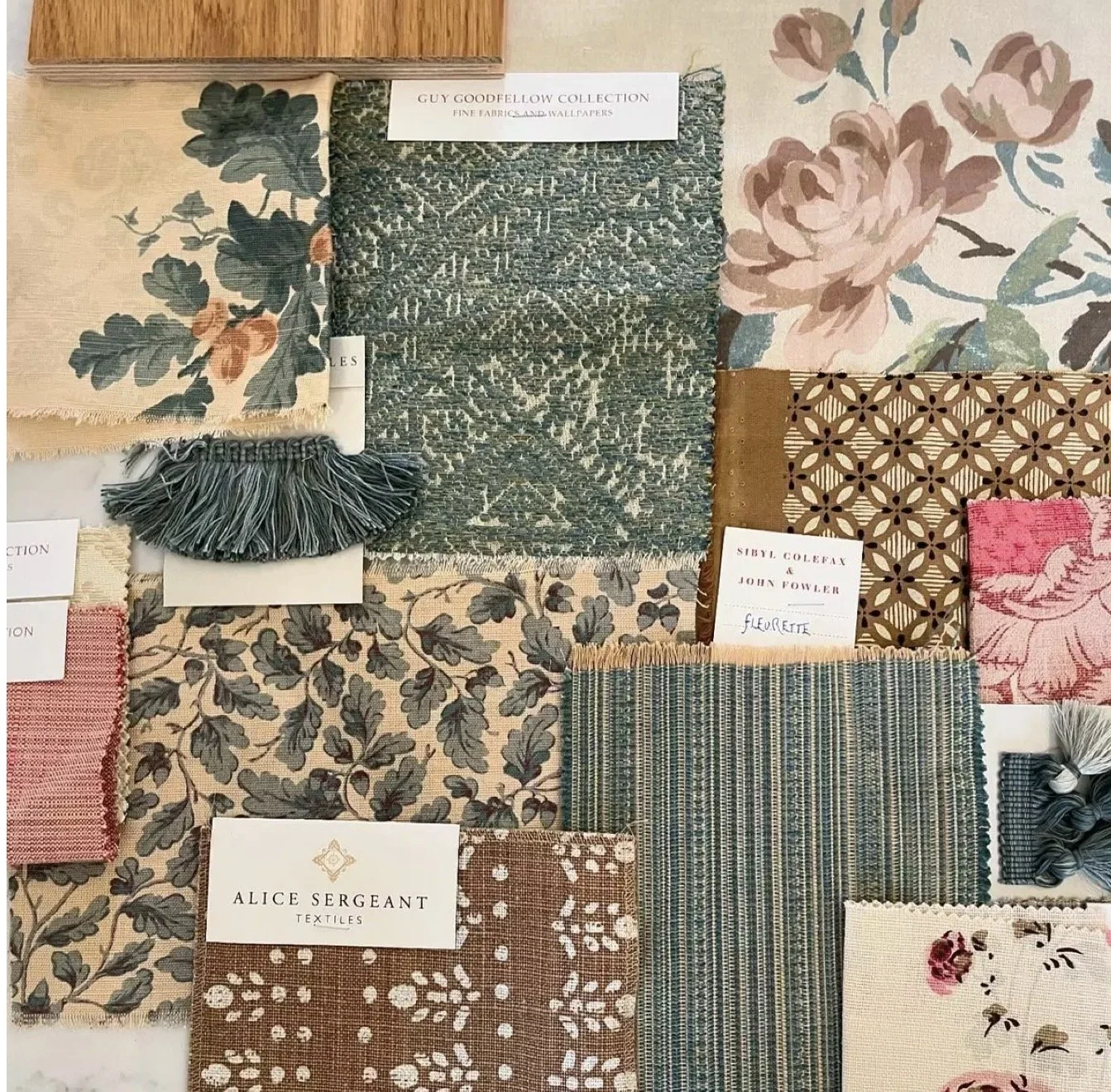 A fabric sample display featuring various textiles, wallpaper, and small fabric swatches with floral and geometric patterns, labeled with designer names.