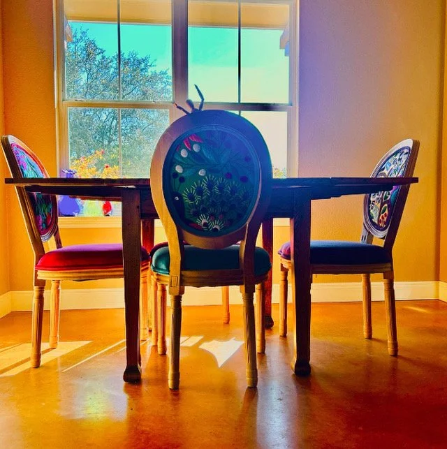 A dining room with a round wooden table and four colorful chairs, sitting in front of a window with sunlight shining through and greenery visible outside.