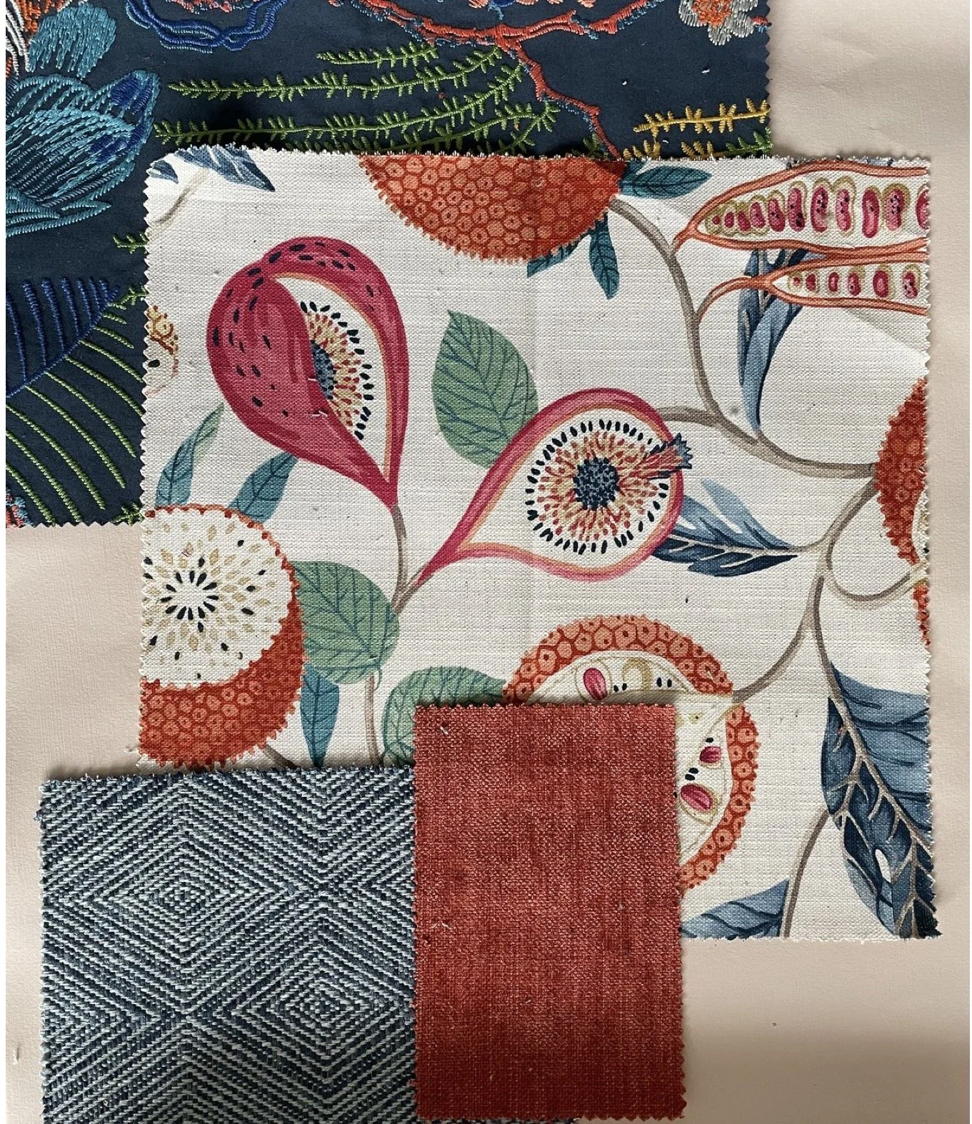 A colorful fabric pattern with floral designs, including large pink and white flowers with black centers, surrounded by green leaves, on a white background. Three smaller fabric swatches in different textures and colors are placed in front of the pat