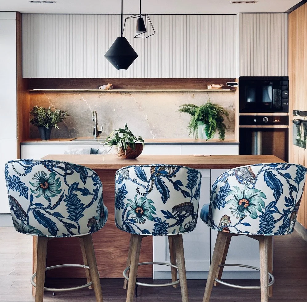 Modern kitchen with a wooden island and three barstools with floral-patterned upholstery. The kitchen features a marble backsplash, potted plants, and built-in black appliances.
