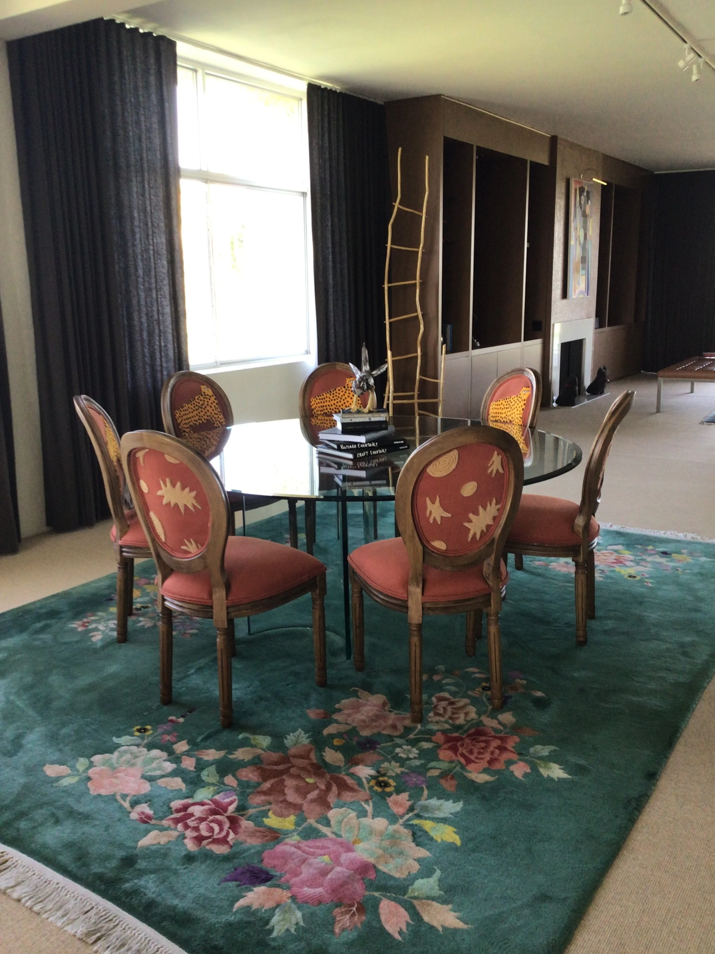 A dining room featuring a round glass table surrounded by six upholstered chairs with coral and yellow patterned fabric. The room has large windows with dark curtains, a floral area rug, and a built-in wooden shelving unit on the wall. There are books and decorative items on the table and a large colorful painting on the wall.