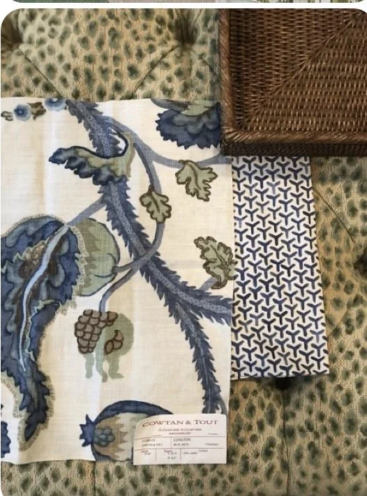 A fabric swatch with a floral pattern in shades of blue, green, and beige, with a small label at the bottom, placed on a patterned green surface with a woven wicker box or tray partially visible in the top right corner.
