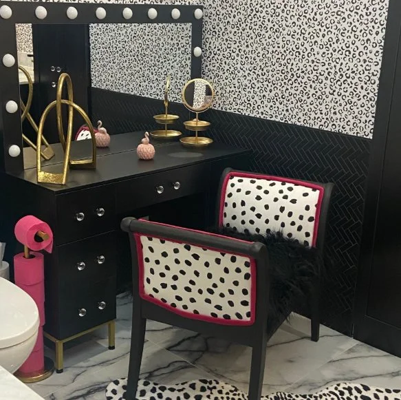 A black vanity table with a mirror, gold decor, and toilet paper held by a pink holder in a black and white themed bathroom with animal print wallpaper and furniture.