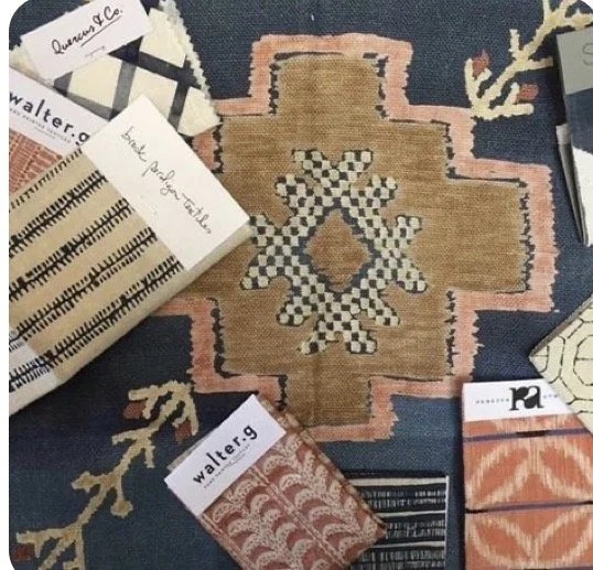 Close-up of a patterned textile with various tags and labels around it, including a large central rug with geometric designs in pink, beige, black, and white, and surrounding smaller fabric samples.