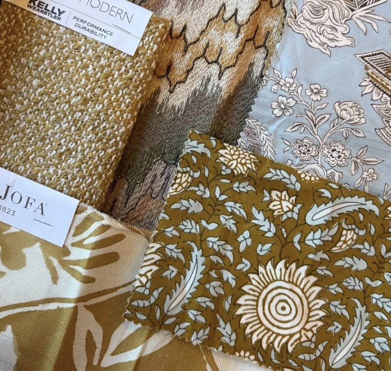 Close-up of various fabric samples with patterns and textures, some with tags indicating names like 'MODERN' and 'JOFA.' The fabrics feature different designs including floral and geometric patterns in shades of brown, beige, white, and blue.