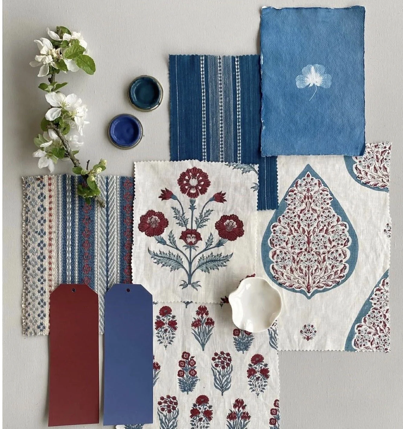 A flat lay of fabric swatches, floral print, paint chips, and dishes, with a branch of white flowers on a light gray background.