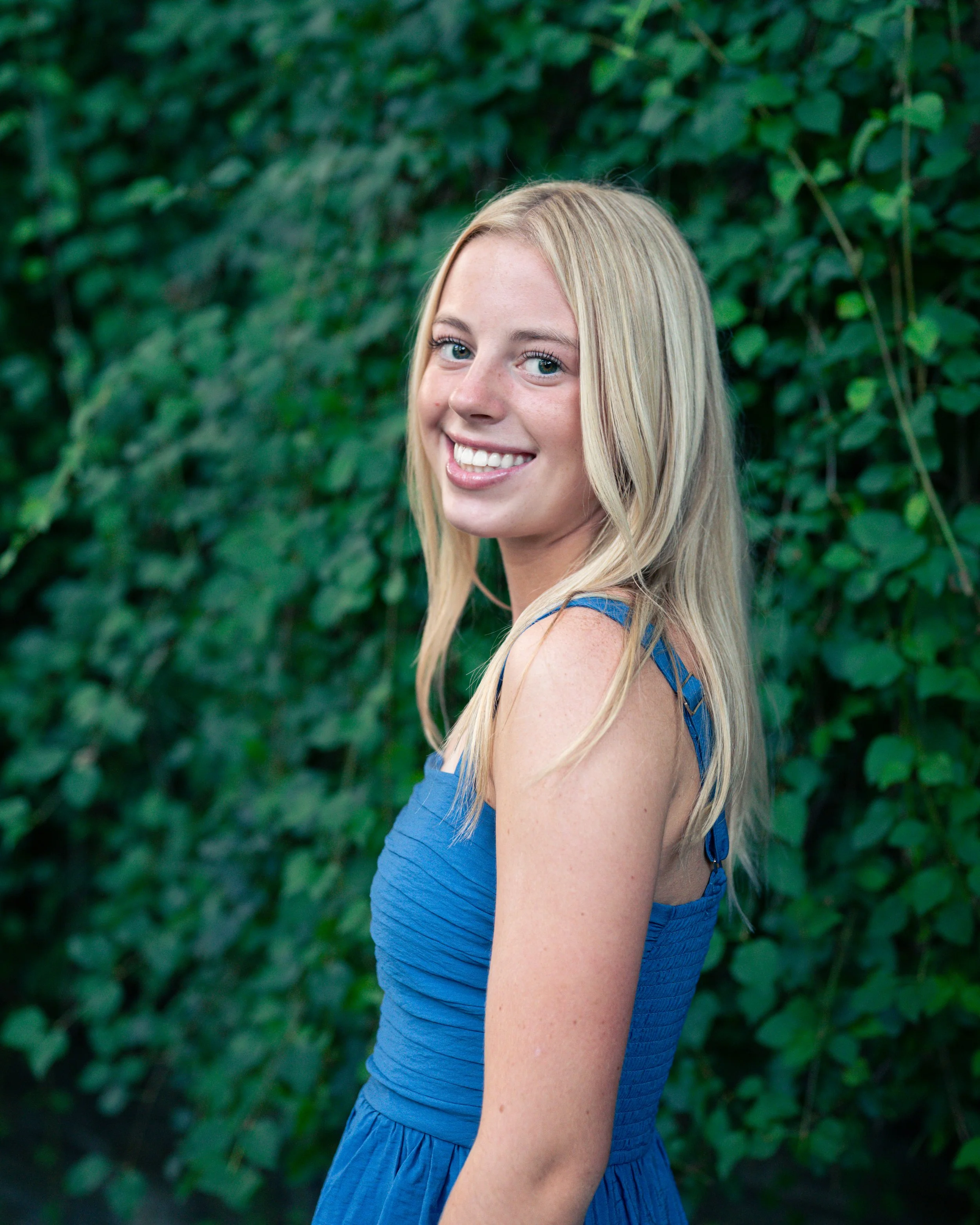 A young woman with long blonde hair and blue eyes smiling outdoors in front of green foliage, wearing a blue sleeveless dress.