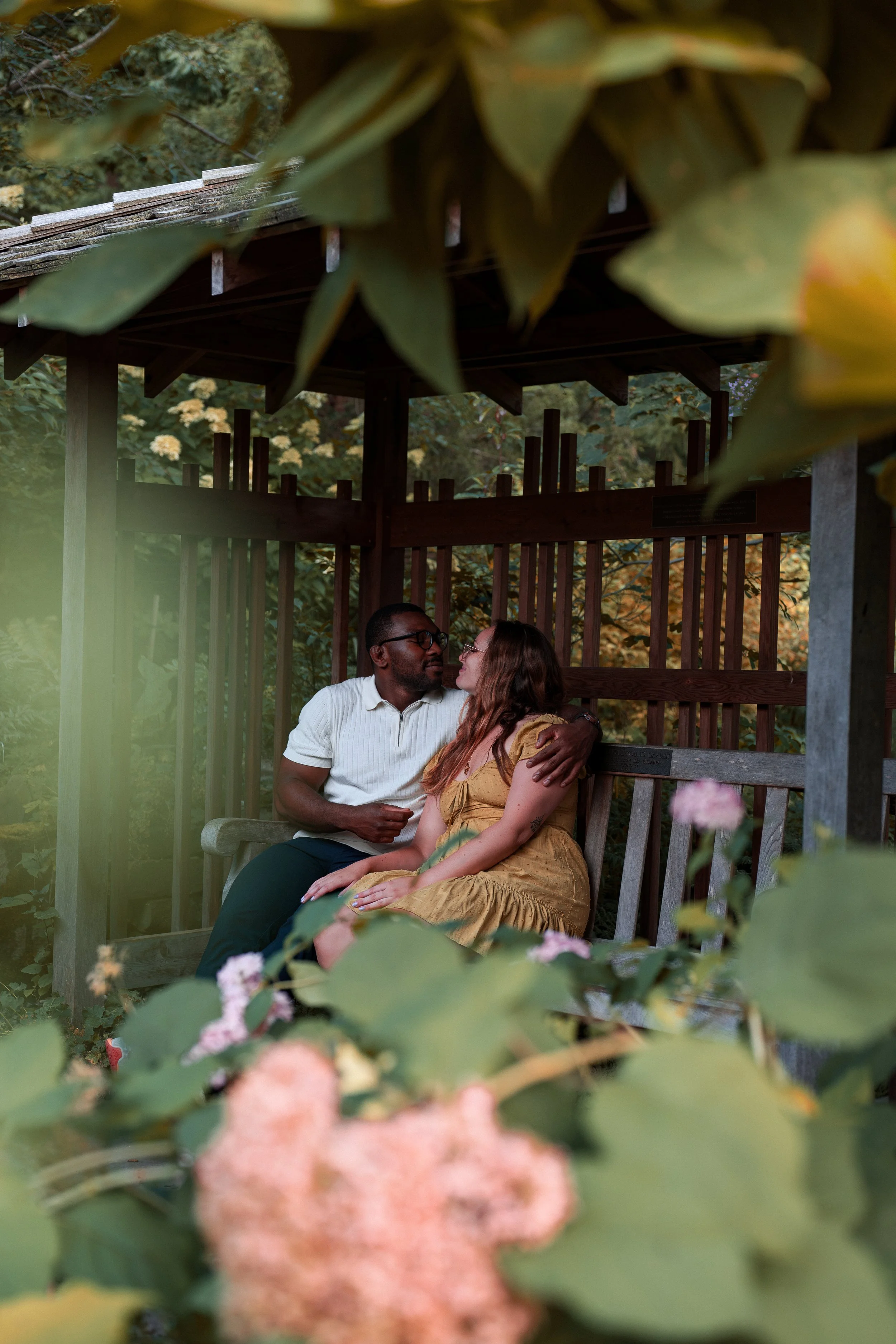 A couple sits on a wooden bench in a garden, surrounded by leafy plants and pink flowers. The man has dark skin, wears glasses and a white shirt, and the woman has light skin, long wavy hair, and a yellow dress. They are looking at each other lovingl