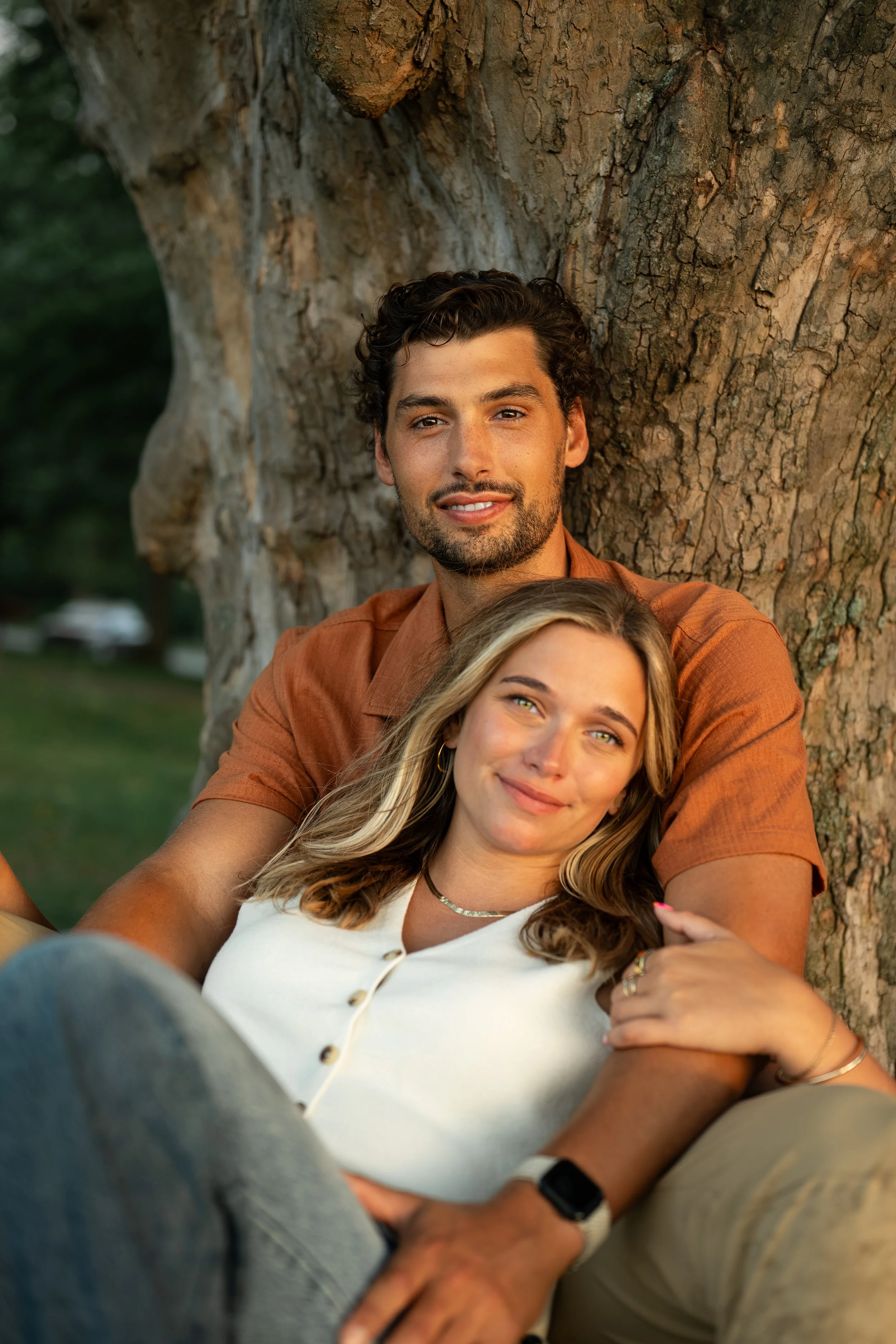 A young man and woman are sitting close together outdoors, leaning against a large tree, enjoying a moment of affection and companionship.