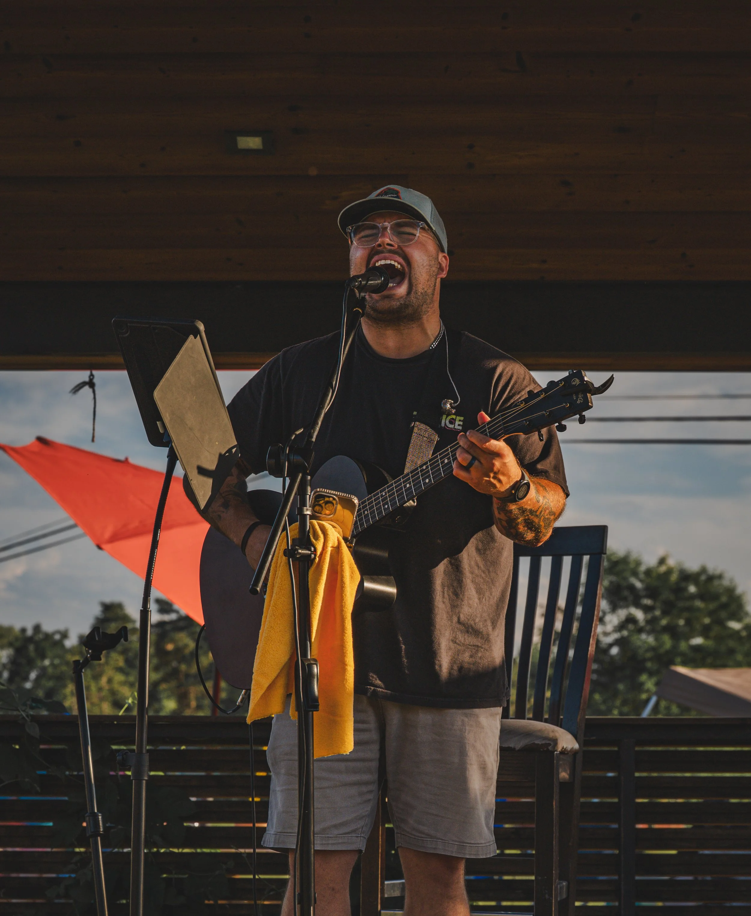 A man singing and playing guitar on an outdoor stage with a dark wooden overhang, wearing a cap, glasses, and a black shirt, with a red umbrella and trees in the background.
