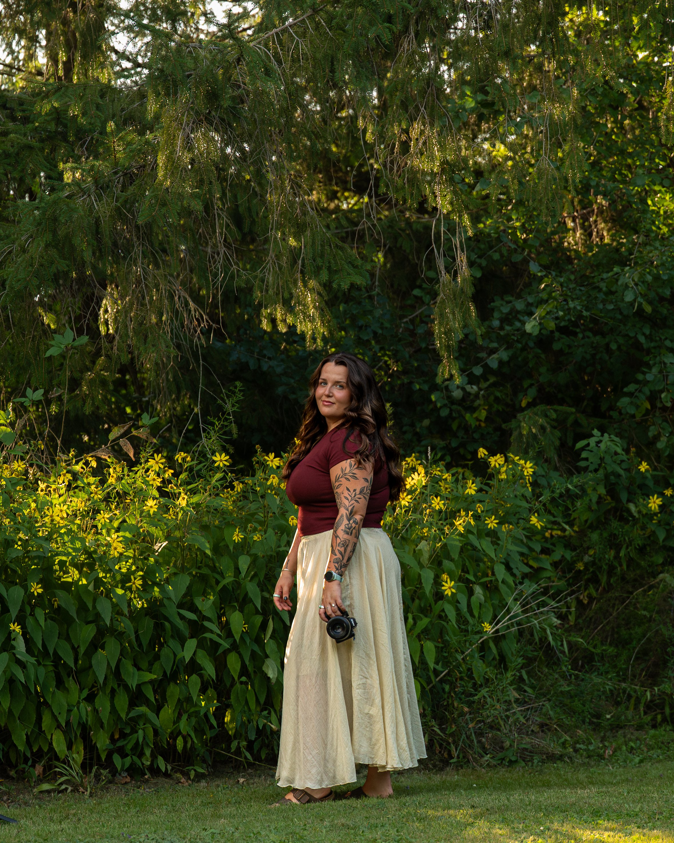 A woman in a long beige skirt and maroon top standing outdoors among greenery and yellow flowers, holding a camera.