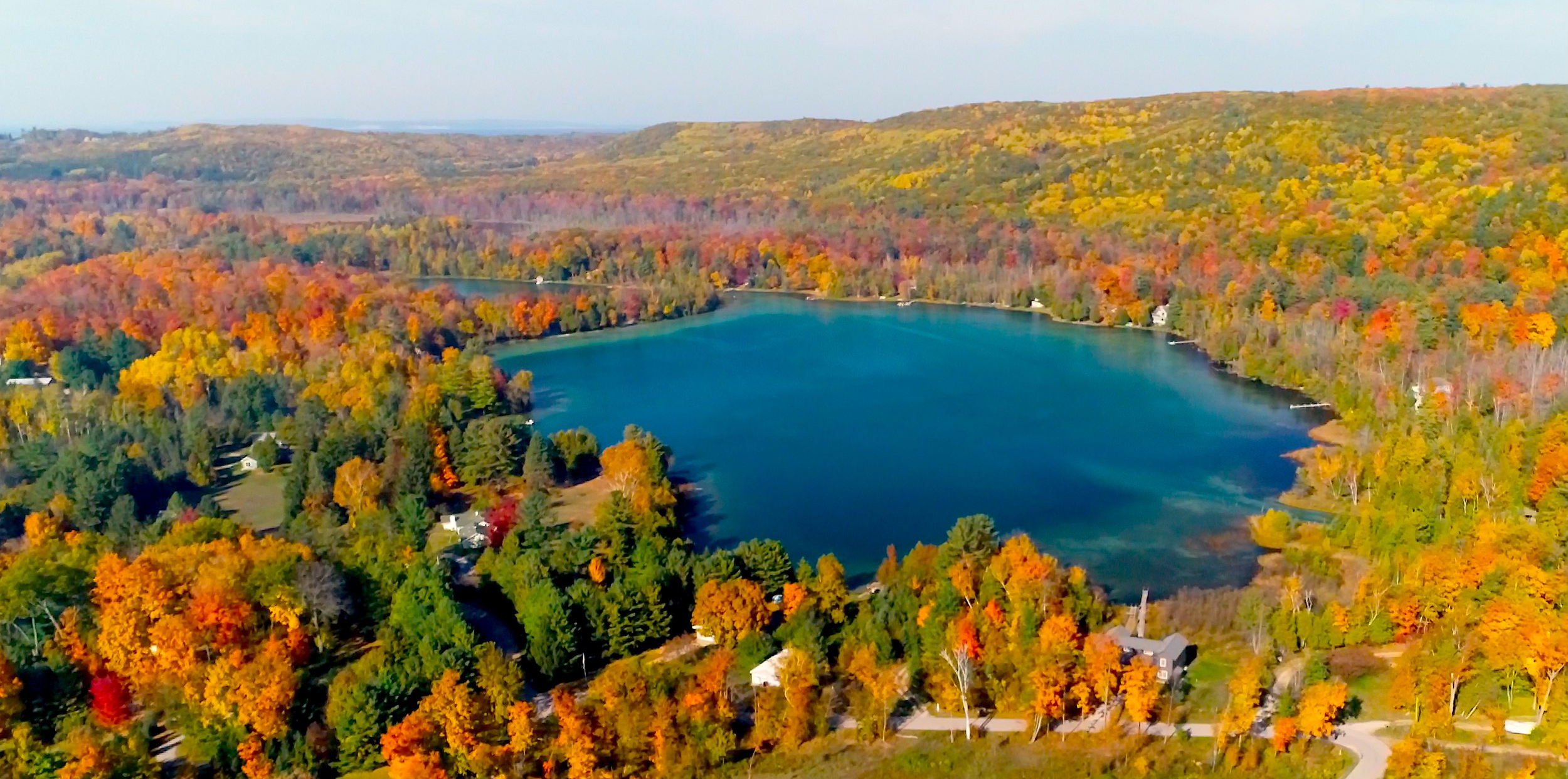 An aerial view of a lake surrounded by colorful autumn trees with houses along the shoreline and rolling hills in the background.