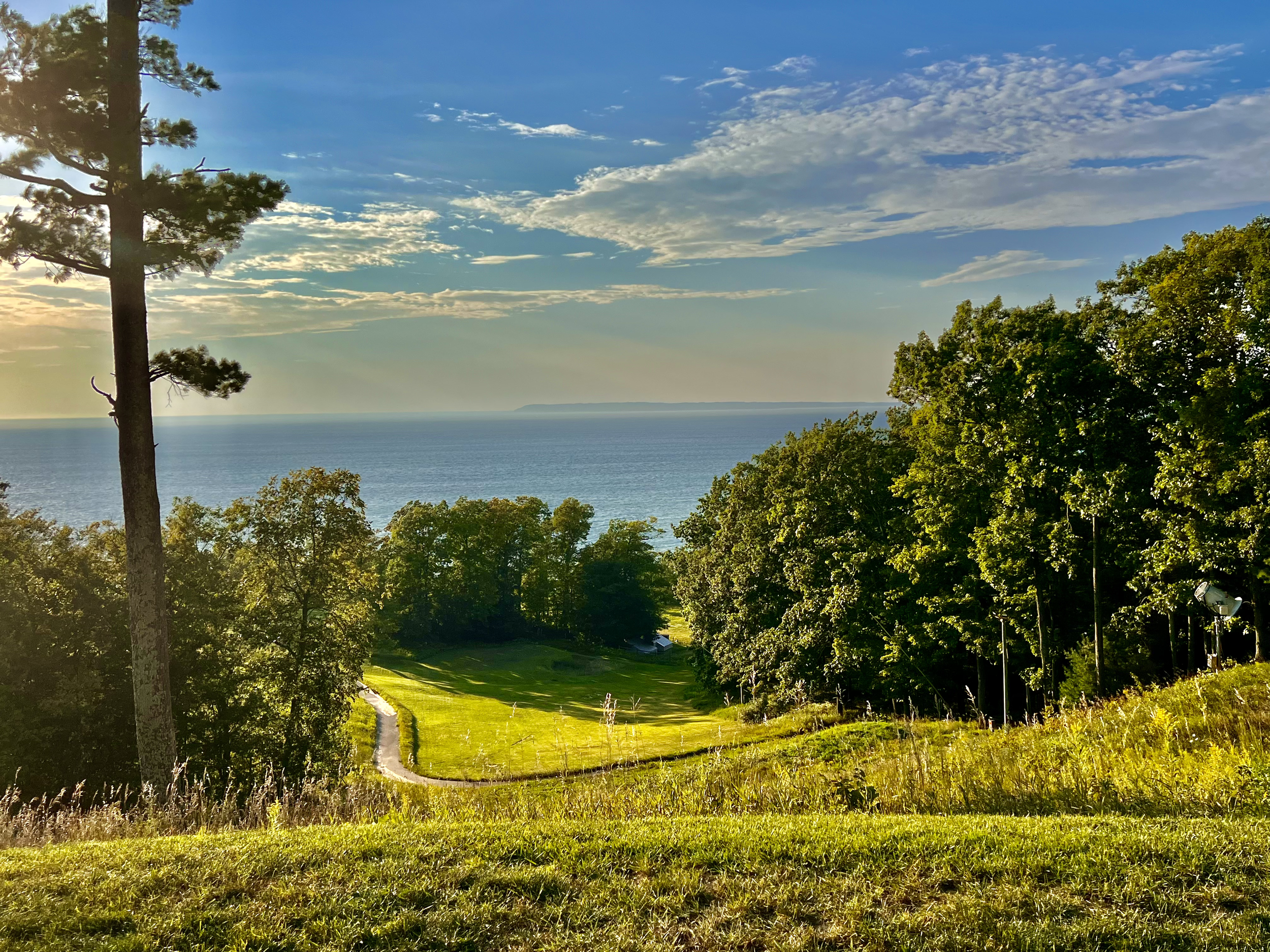 Scenic view of a golf course near a body of water, surrounded by trees, under a partly cloudy sky with the sun setting.